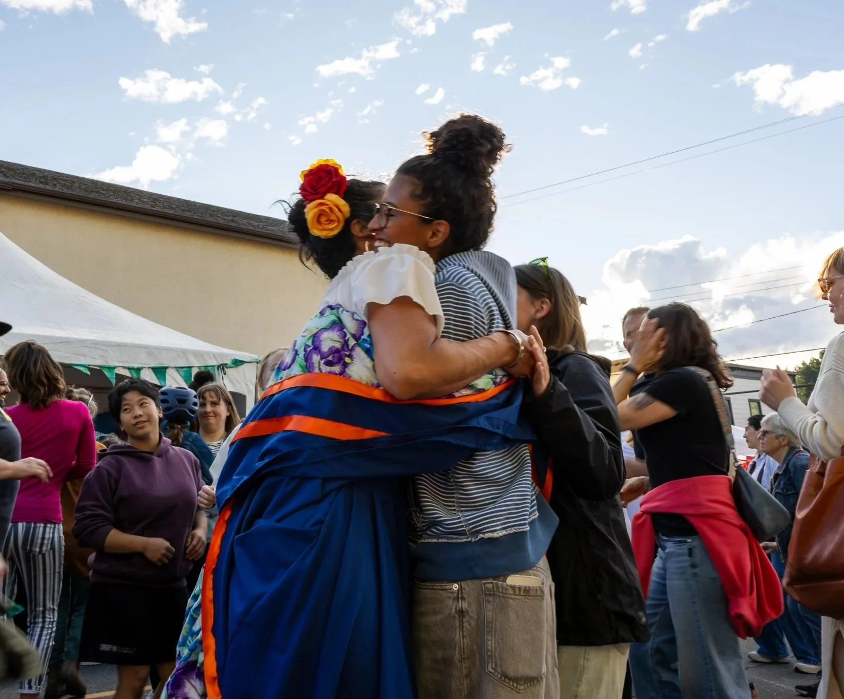 Two women hugging and smiling at each other during an outdoor event, with a crowd of people in the background including children and adults, some wearing glasses, and a white tent on the left, under a partly cloudy sky.