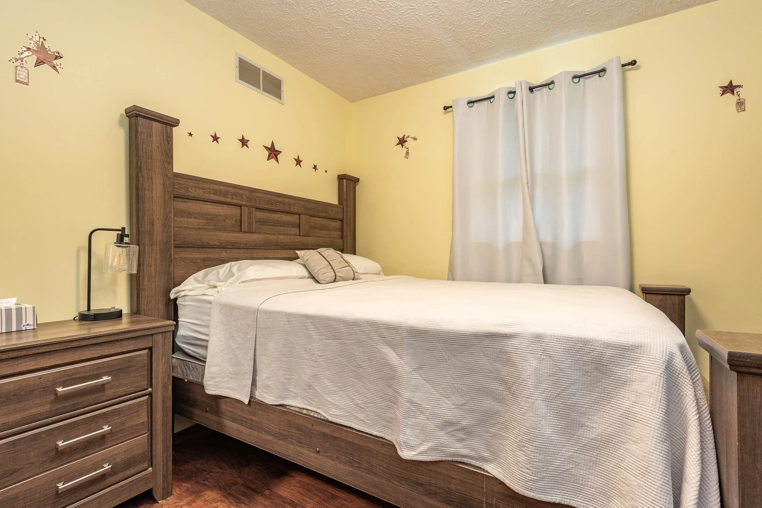 A bedroom with a wooden bed frame, a white bedspread, a beige pillow, a nightstand with a black lamp, and yellow walls decorated with red star and gift stickers. White curtains hang over a window. There is a vent in the ceiling.