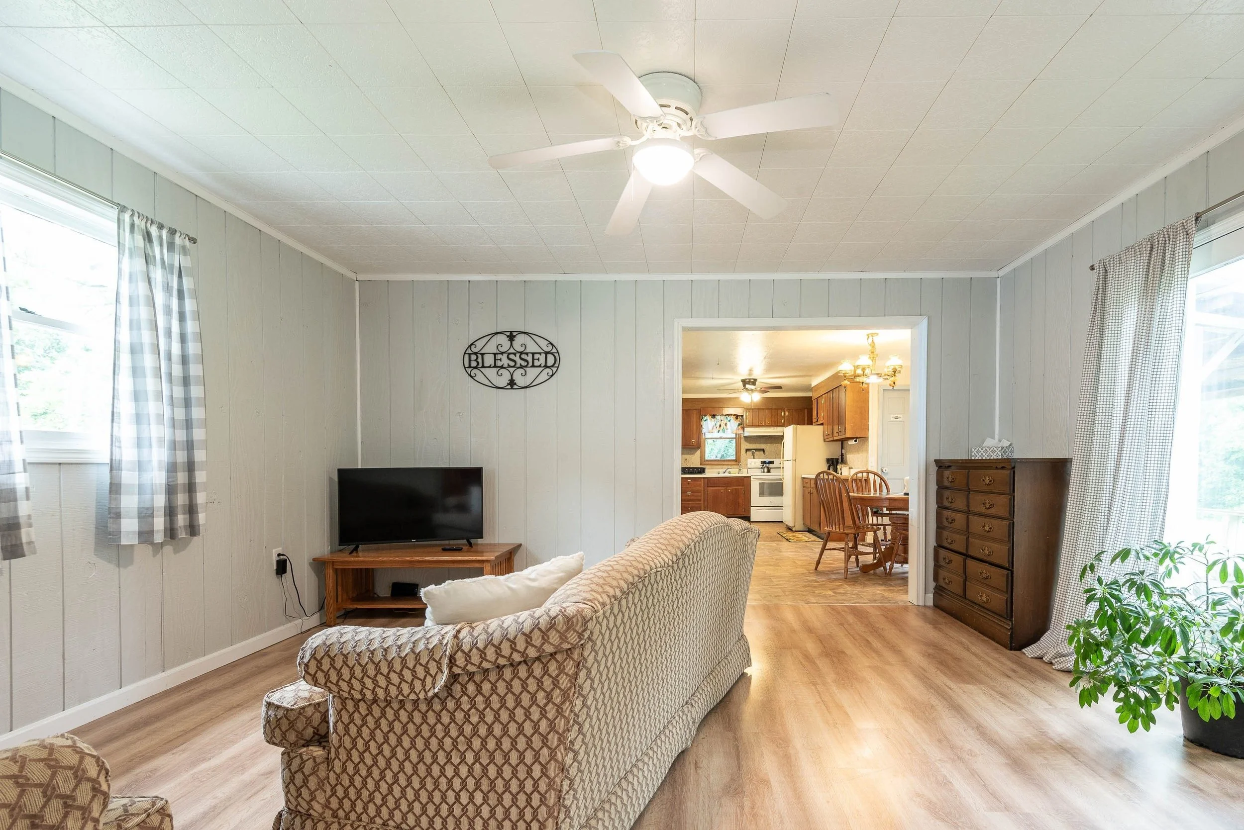 Living room with beige patterned sofa, wooden coffee table, wall-mounted TV, and an opening to a dining area with wooden furniture and kitchen in the background. Windows with checkered curtains and a ceiling fan with light.