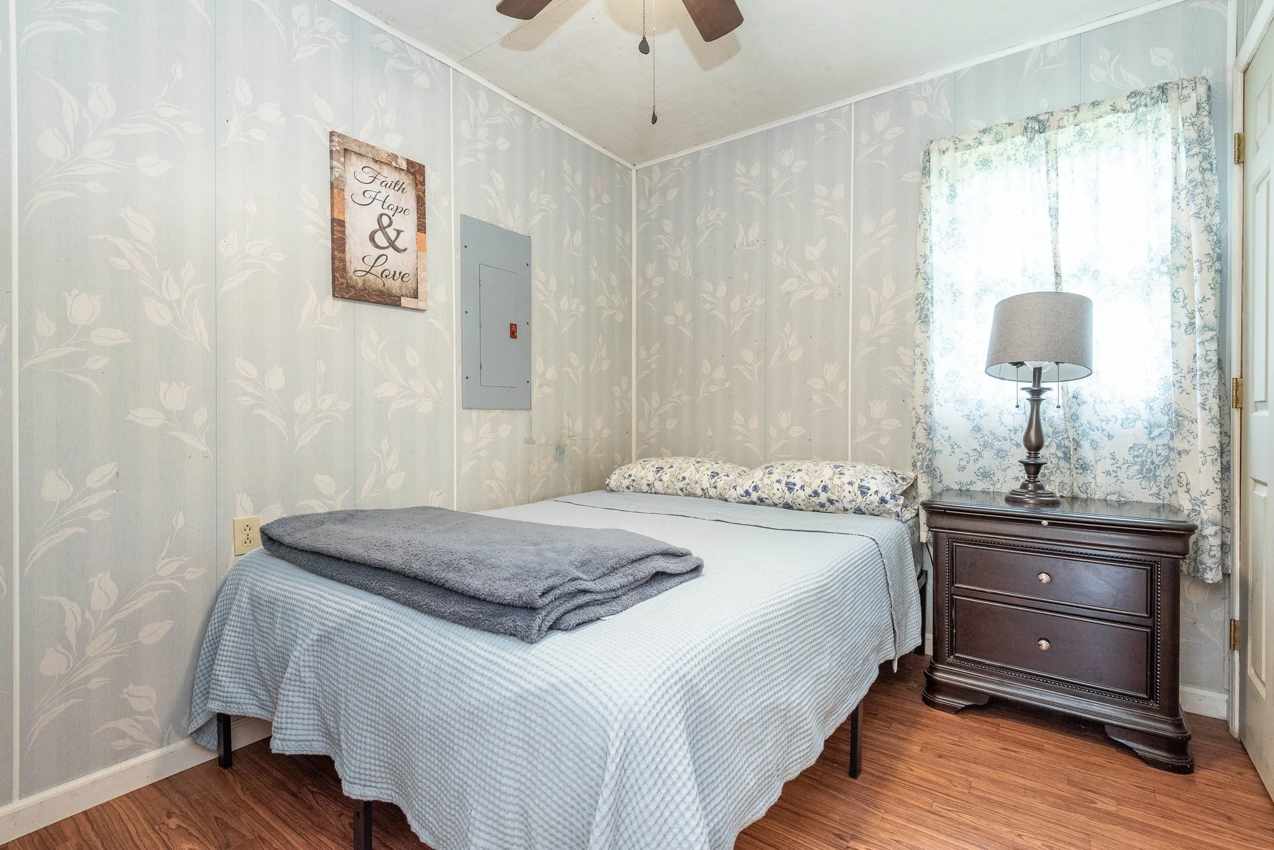 A bedroom with a bed, nightstand, and lamp. The bed has a blue and white checkered blanket, a folded gray towel at the foot, and a floral pillow. There is a window with floral curtains, a wall decoration with the words 'Faith, Hope, & Love,' and a ceiling fan.