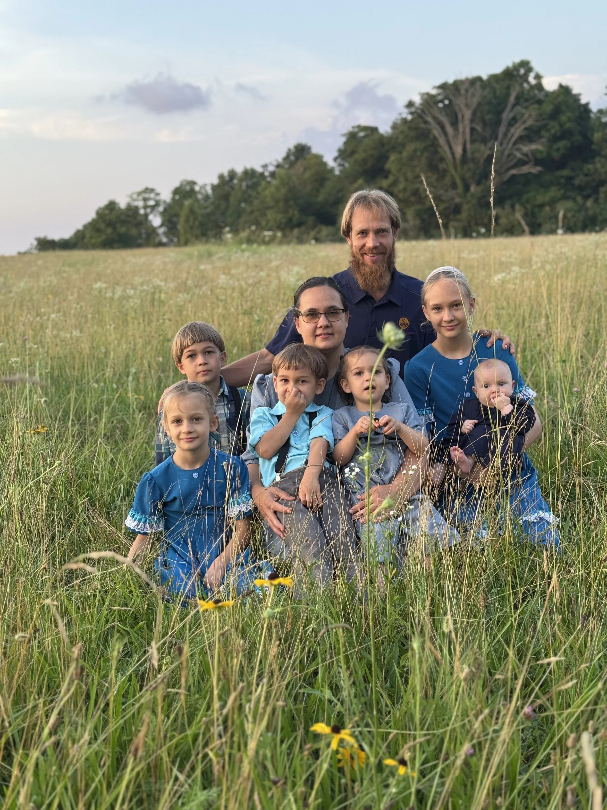 Family photo in a field of tall grass with wildflowers, with trees and a partly cloudy sky in the background.