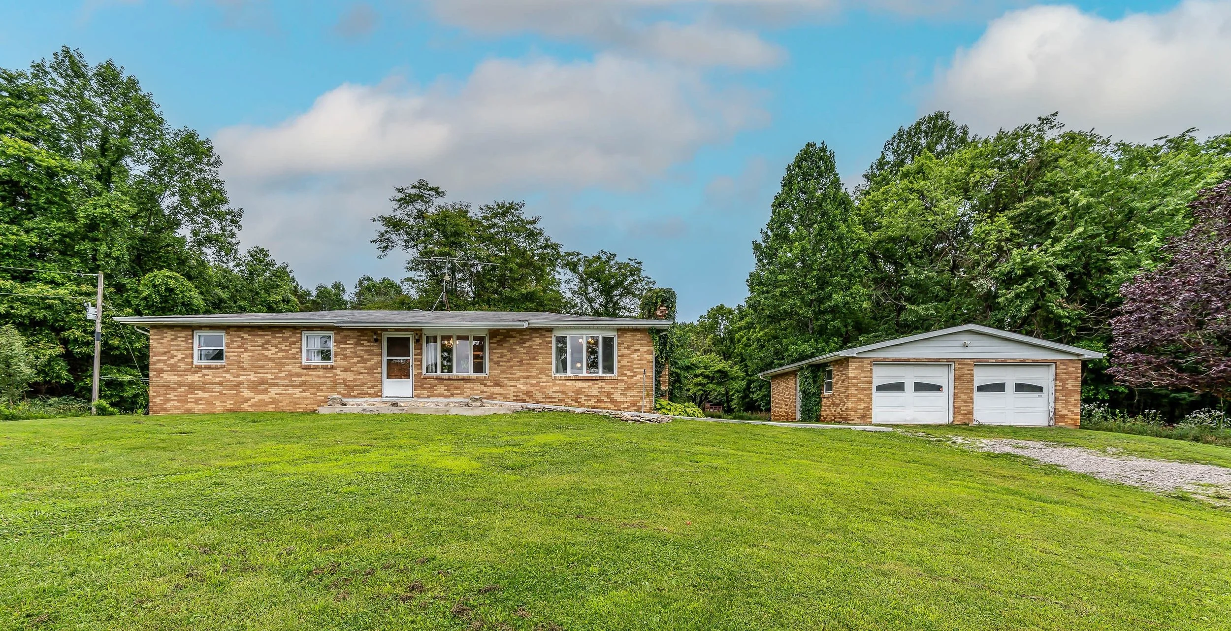 A single-story brick house with a front door, three windows, and a separate two-car garage on a grassy lawn surrounded by trees under a partly cloudy sky.