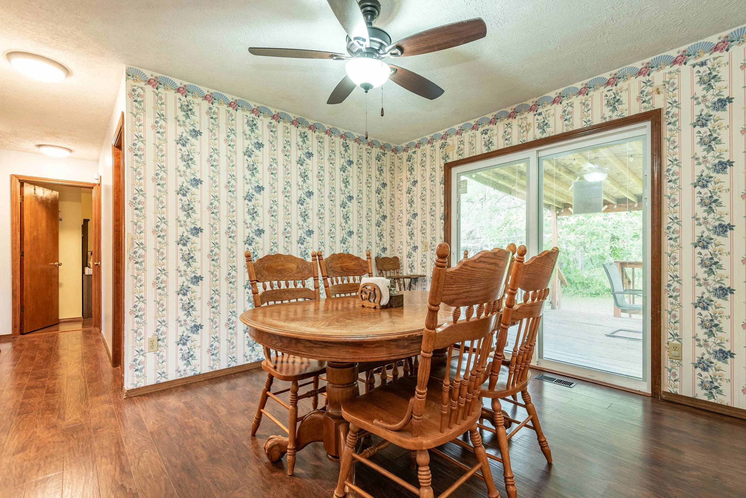 Dining room with a wooden table and chairs, floral wallpaper, a ceiling fan, and a large sliding door leading to an outdoor deck.