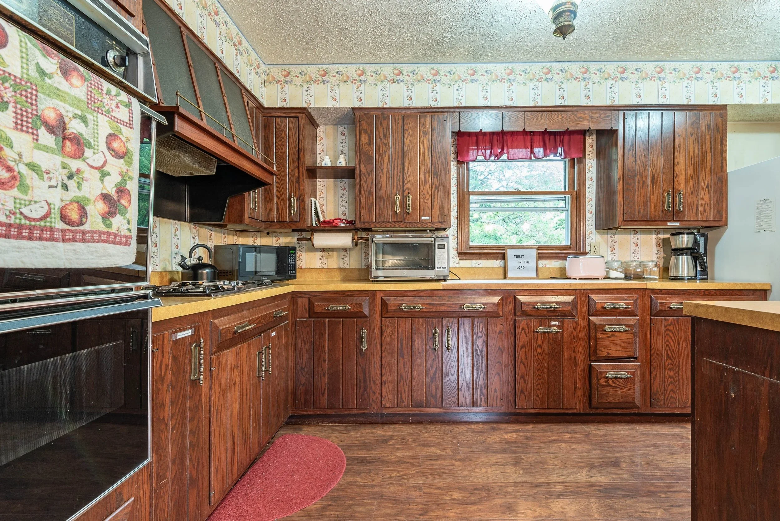 Kitchen with wooden cabinets, a window with red curtains, microwave, toaster, coffee maker, and various kitchen items on the countertops. There is a red rug on the floor and a wallpaper border with fruit motifs on the walls.