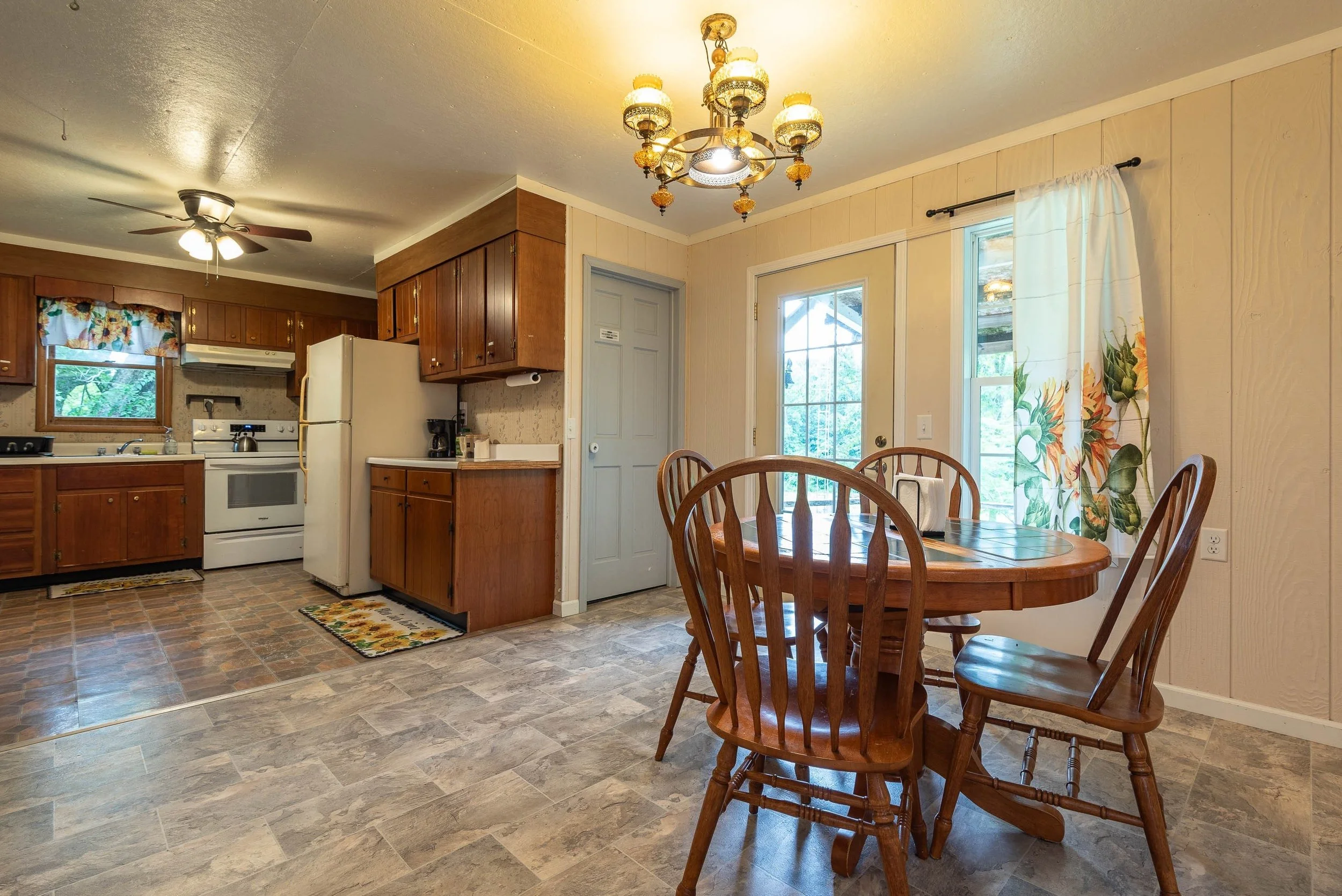 A cozy kitchen and dining area with wooden cabinets, a white stove, and a round wooden dining table with chairs. The room has a window with floral curtains, a glass door, and ceiling lights.