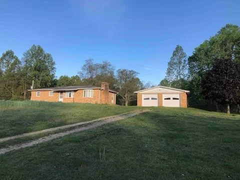 Single-story brick house with a chimney and attached garage, situated on a grassy hill with a dirt driveway, surrounded by trees under a clear blue sky.