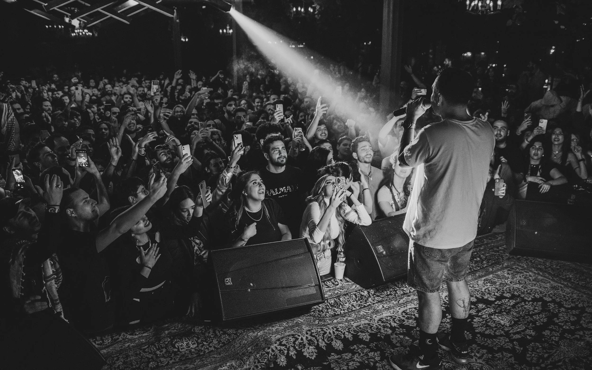 An artist performs on stage with a microphone in front of a large, cheering audience at a concert or music event. The audience is holding up their phones to record or take pictures, and some have their hands raised.