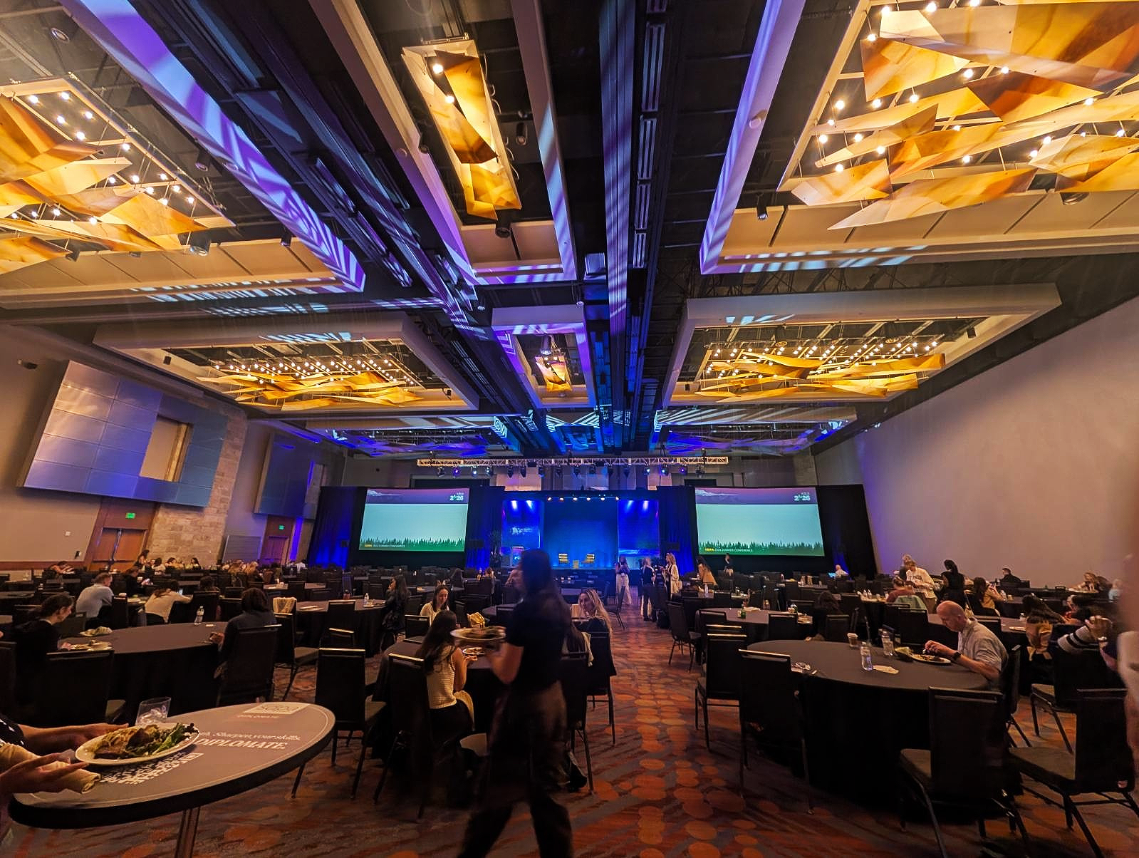 A large conference room filled with round tables and chairs, some people sitting and eating, a stage with two large screens, and sophisticated ceiling decorations with lighting.