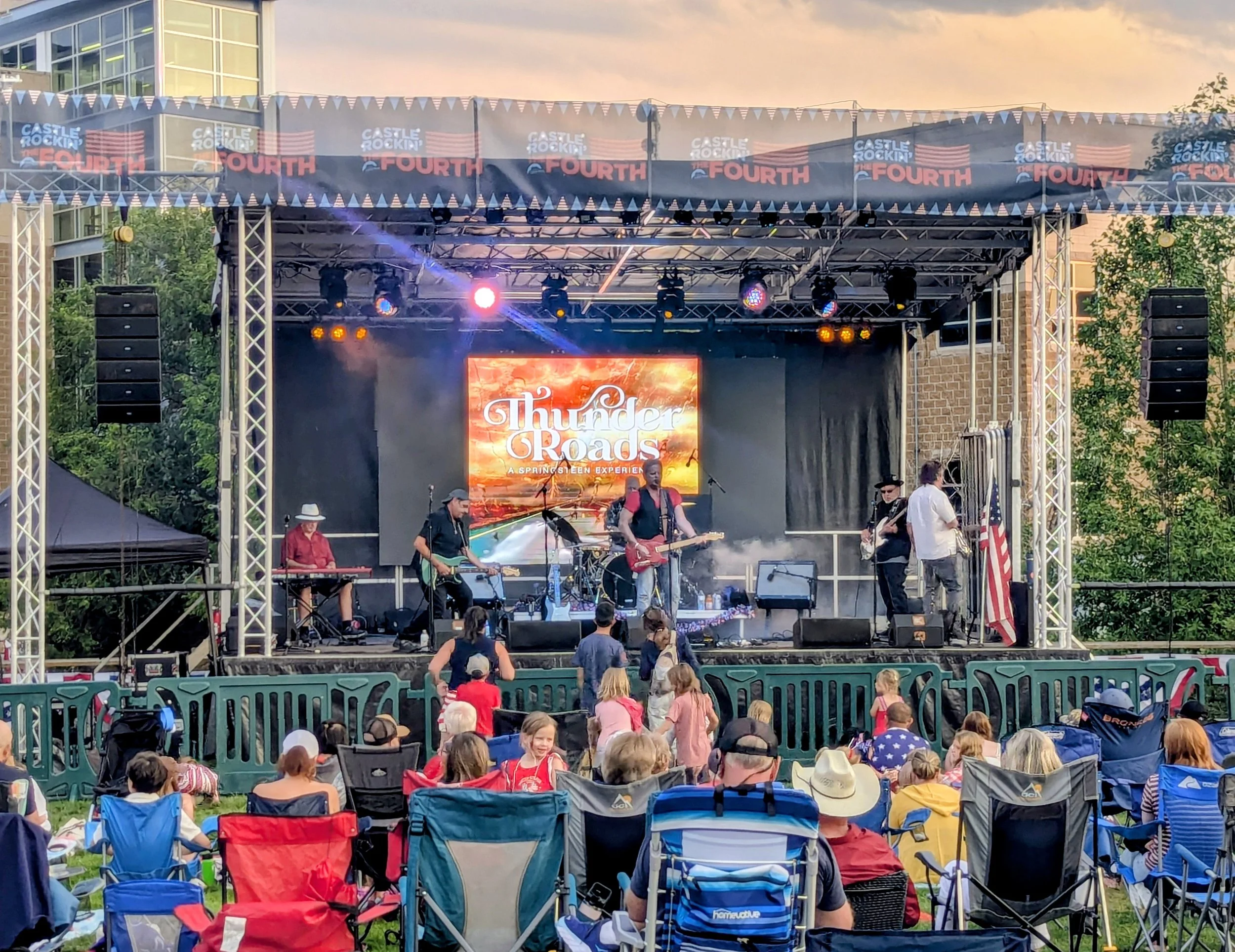 An outdoor concert stage with a live band performing. The stage has LED screens displaying the words 'Thunder Roads' and a smaller caption 'A Springsteen Experience.' Audience members are seated in front of the stage in chairs and on the grass, with 