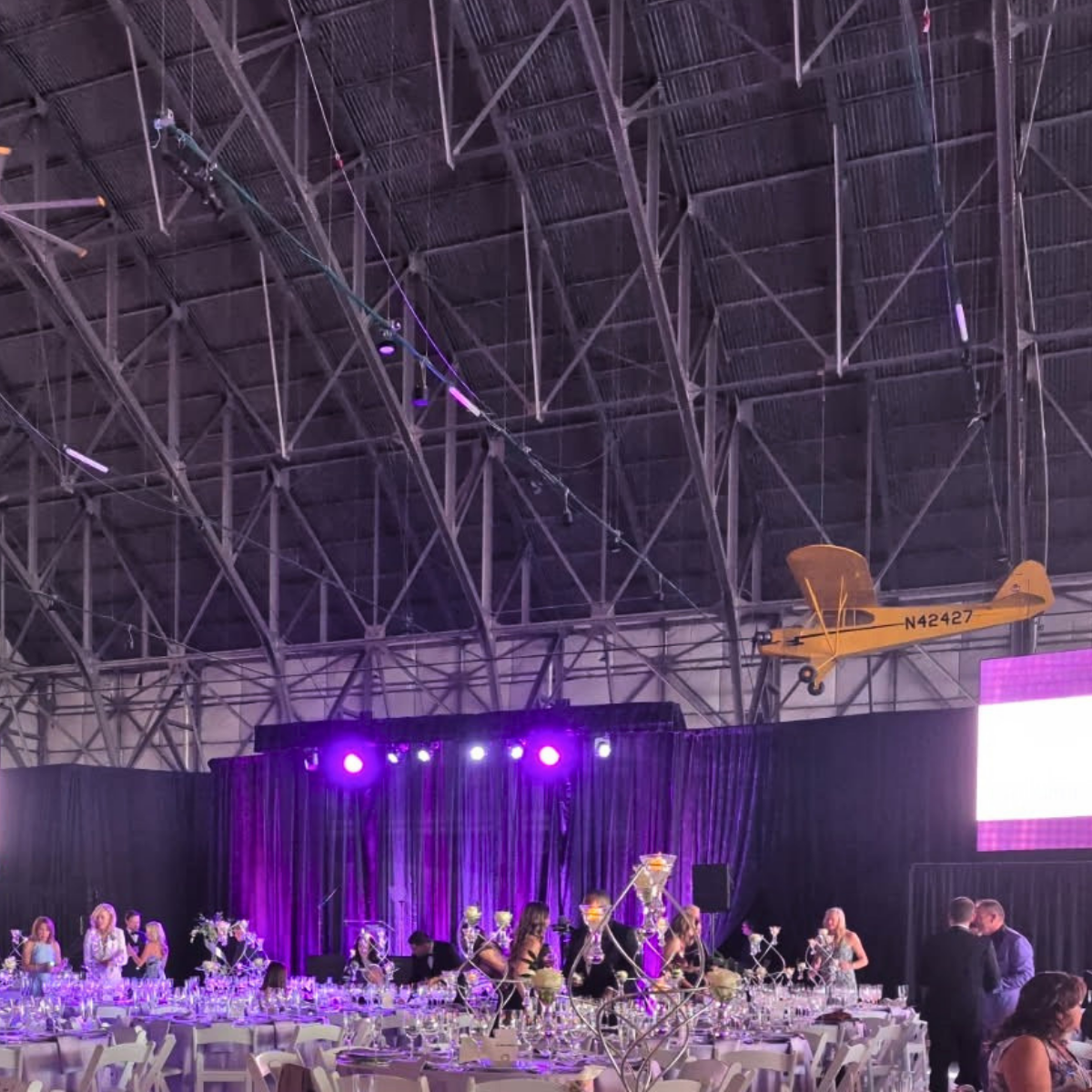 Indoor event space with purple lighting, decorated banquet tables, and a black curtain backdrop. There is a yellow vintage aircraft hanging from the ceiling and a person cleaning high on a cleaning scaffold.