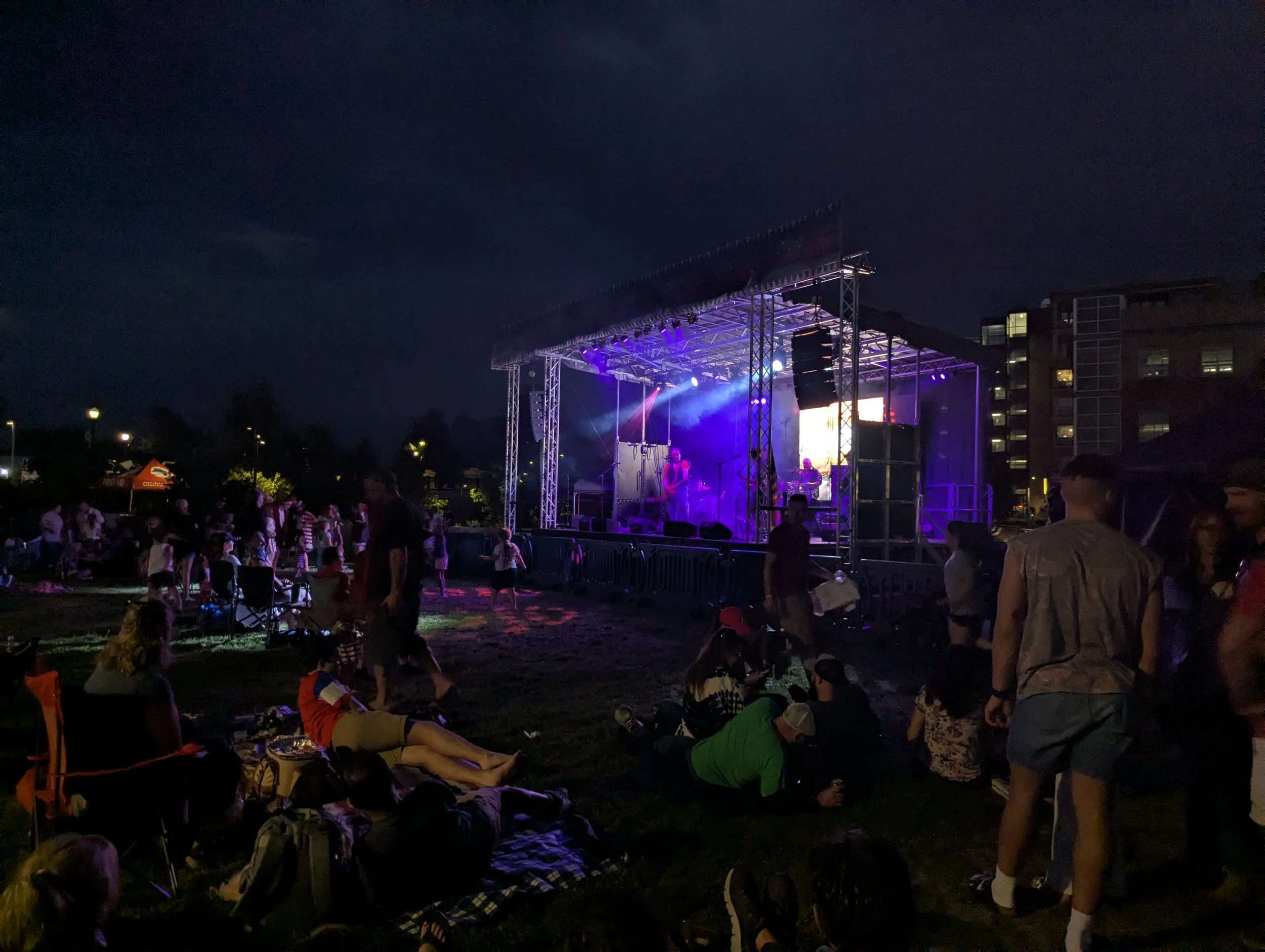 People gathered outdoors at night watching a live concert on a stage with purple and blue lighting.