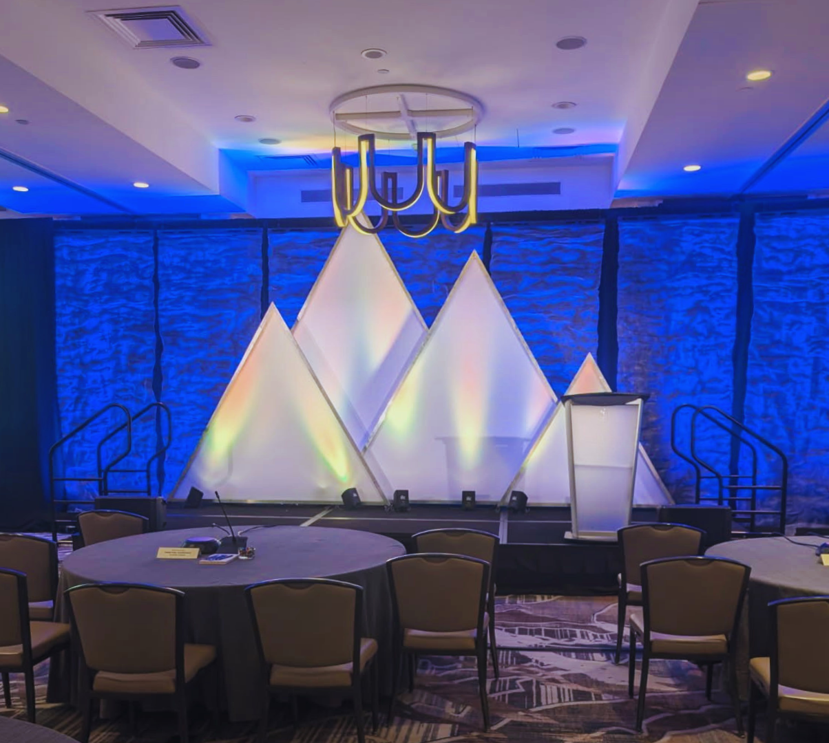 Event stage with geometric white panels illuminated with colorful lights, blue textured backdrop, and a modern chandelier hanging from the ceiling, surrounded by round tables and chairs.