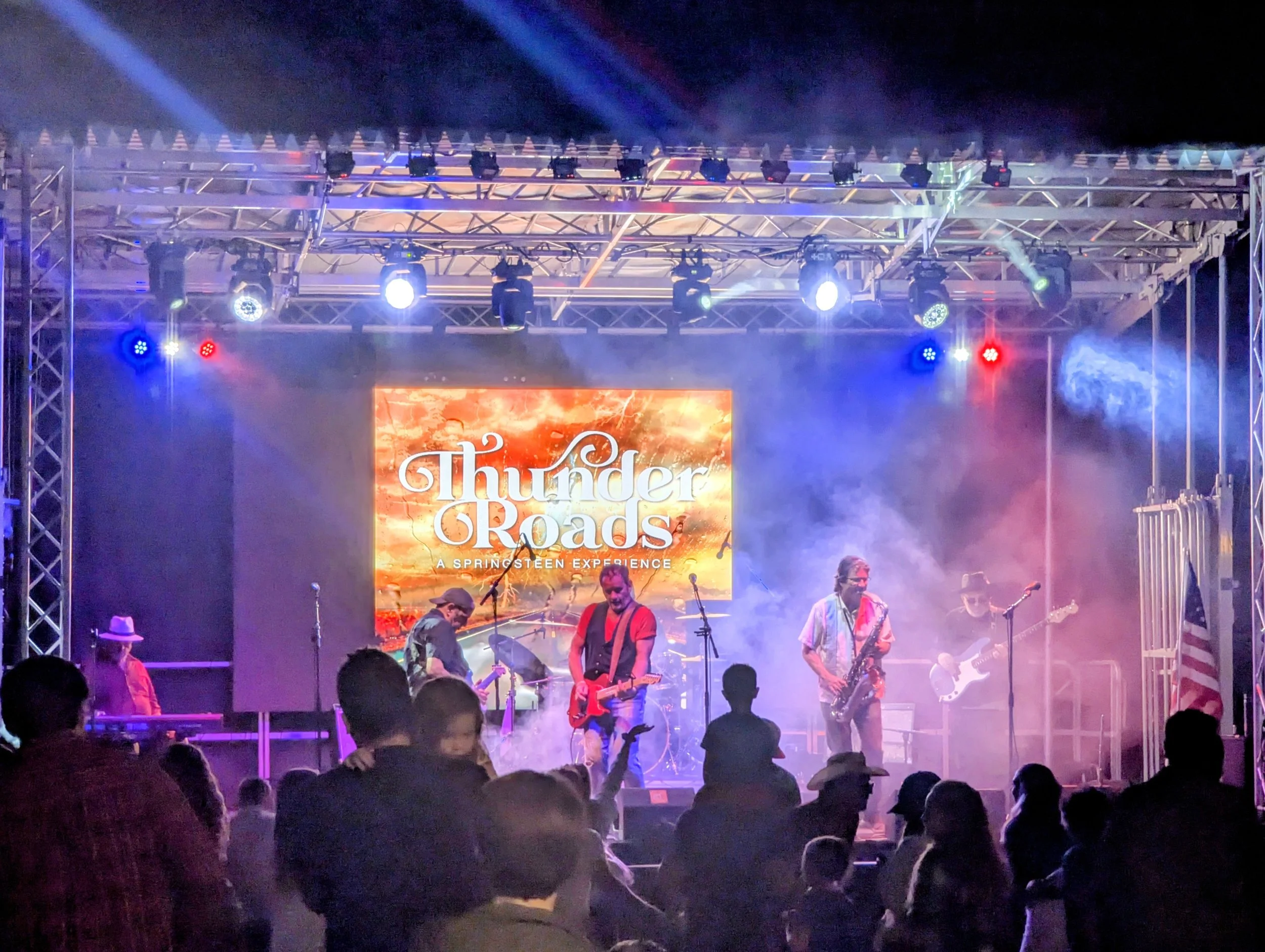 A live concert stage with a band performing under colorful lighting, featuring a large banner that reads "Thunder Roads" and a subtitle "A Springsteen Experience." The audience is visible in the foreground with people watching the performance.