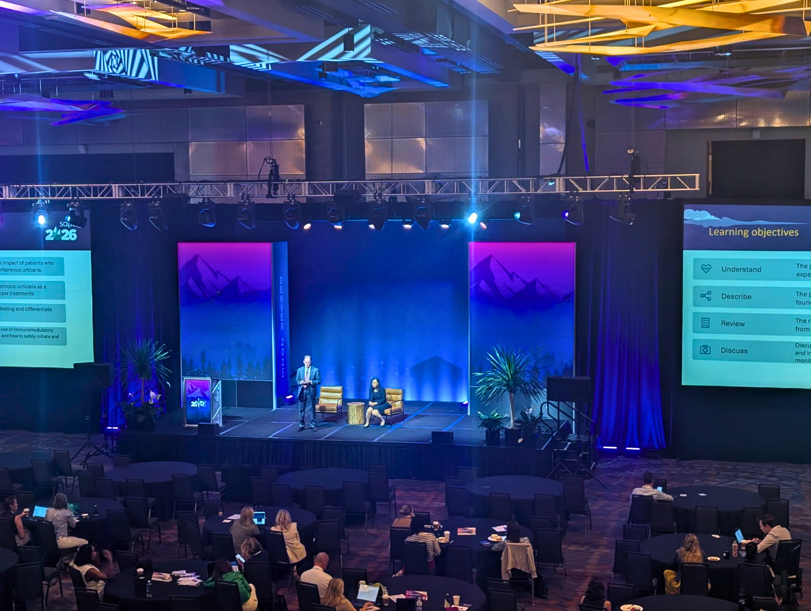 A large conference hall with a stage at the front, where a presentation is taking place. The stage has two speakers, one standing and one seated, with presentation slides projected on screens to the sides. The background features mountain imagery and