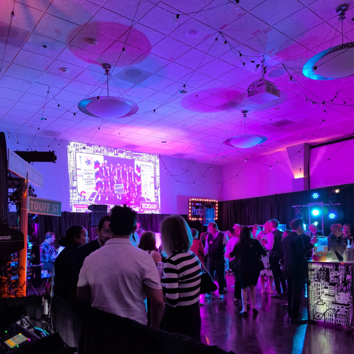 Indoor event with purple and blue lighting, people gathered around, a large screen displaying a group photo, and various banners and signs.