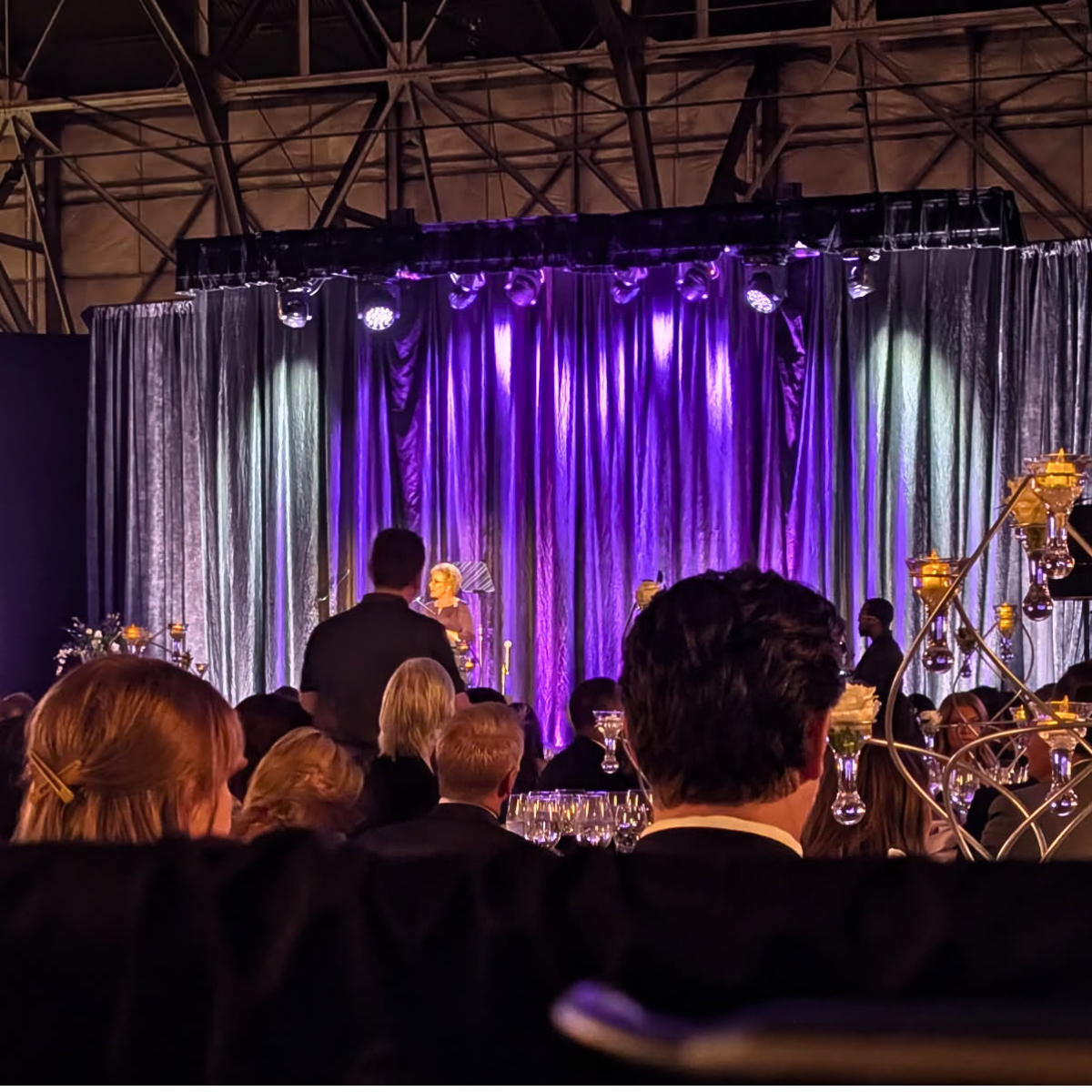 An indoor event with a stage and purple curtains. People seated at tables, some with glasses and floral centerpieces. A woman appears to be speaking or performing on stage, with others standing nearby. The lighting is warm and elegant, with decorativ
