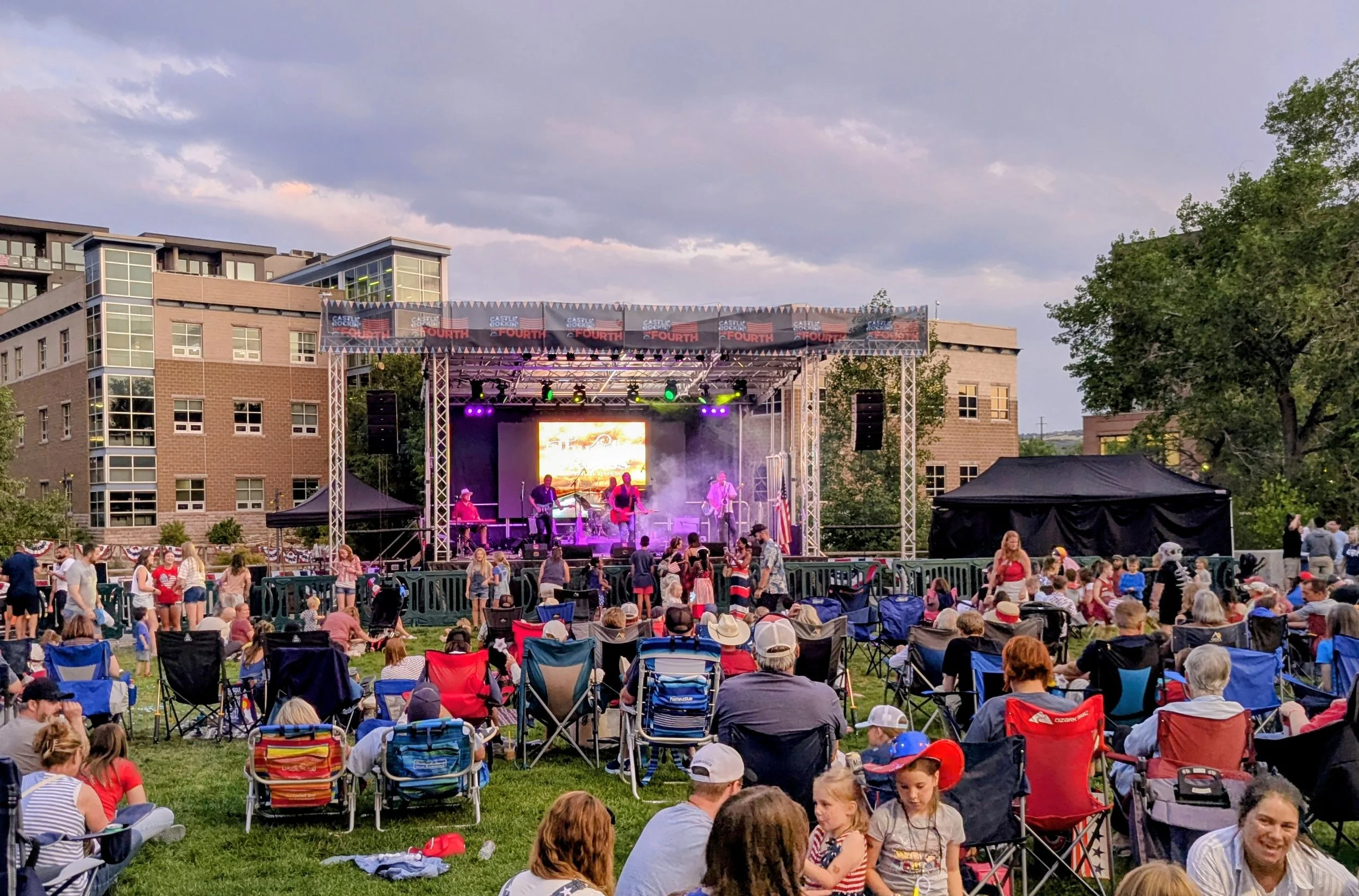 A crowd of people watching a live music performance outdoors on a stage, with some seated in lawn chairs and others standing. The stage has colorful lights and a large screen, with buildings and trees visible in the background under a cloudy sky.