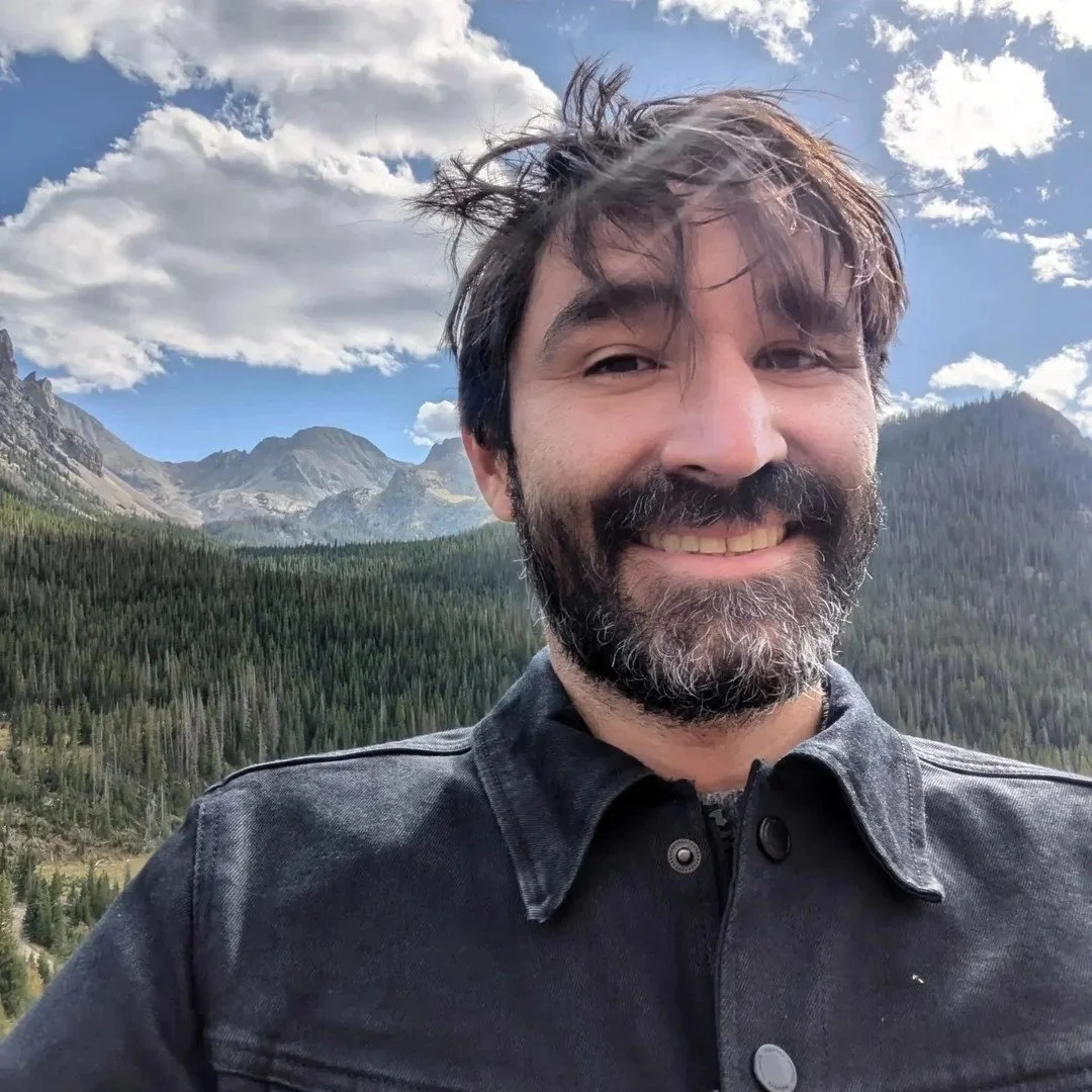 A smiling man with dark hair and beard taking a selfie with mountain scenery and trees in the background under a partly cloudy sky.