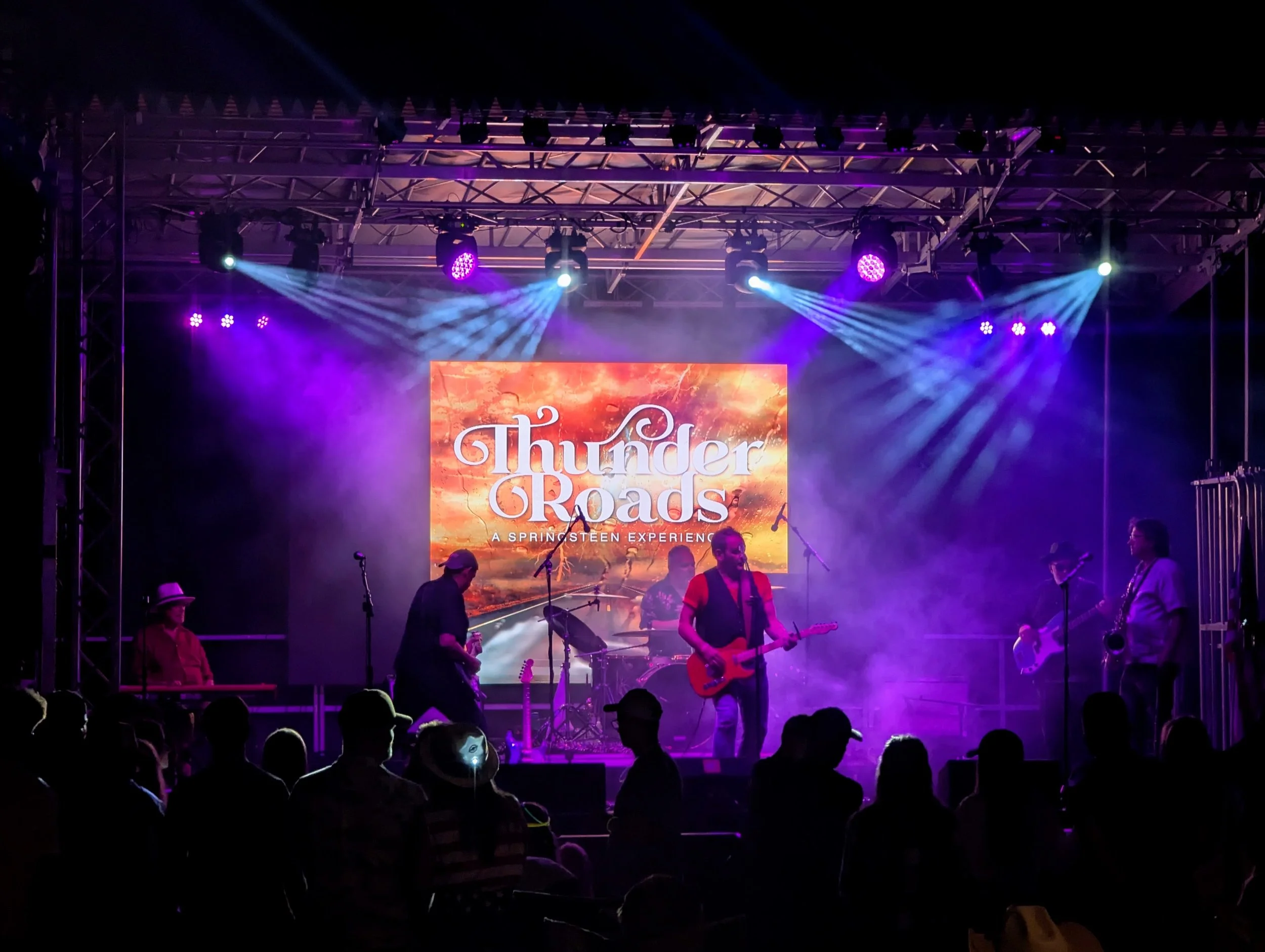 A band performing on stage at a concert with bright colorful lights and a large screen displaying the words "Thunder Roads" and "A Springsteen Experience." Audience members are visible in the foreground.