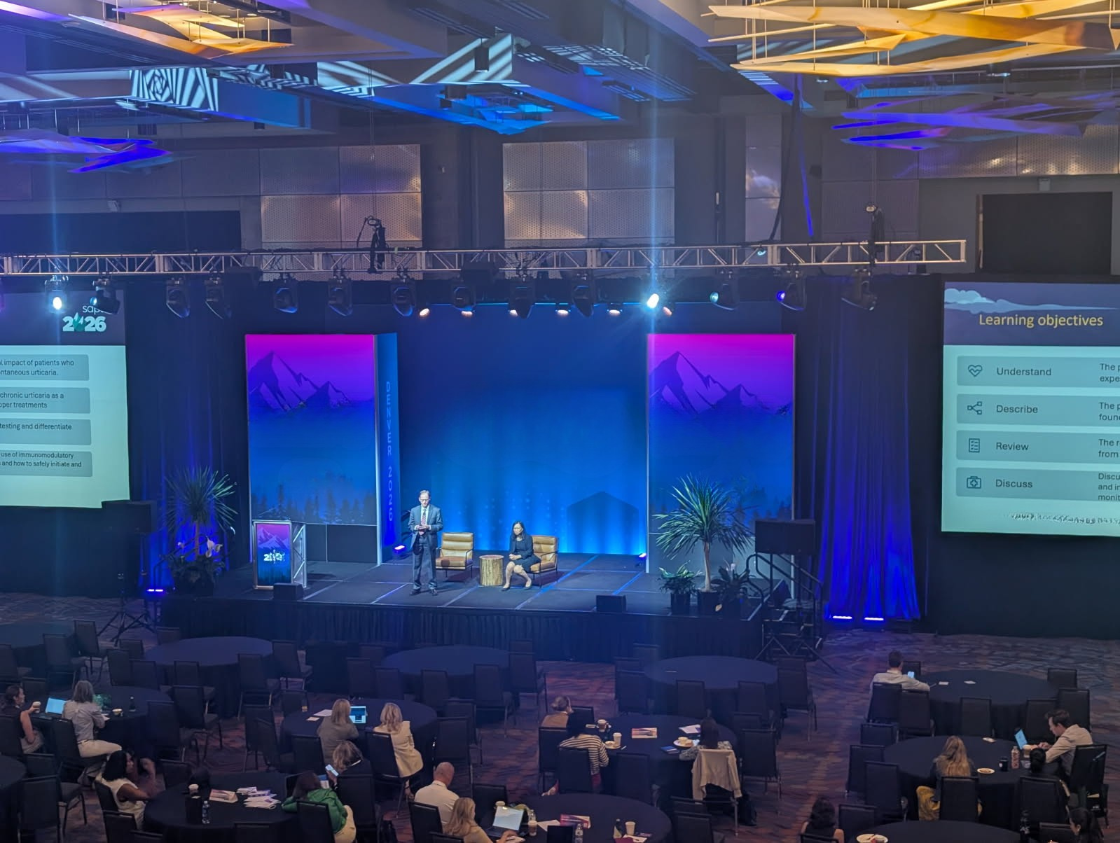 A conference stage with a presenter speaking into a microphone, and a seated panelist on a chair. Two large screens display presentation slides, with the right slide titled 'Learning objectives'. The stage has plants and is illuminated with colorful 