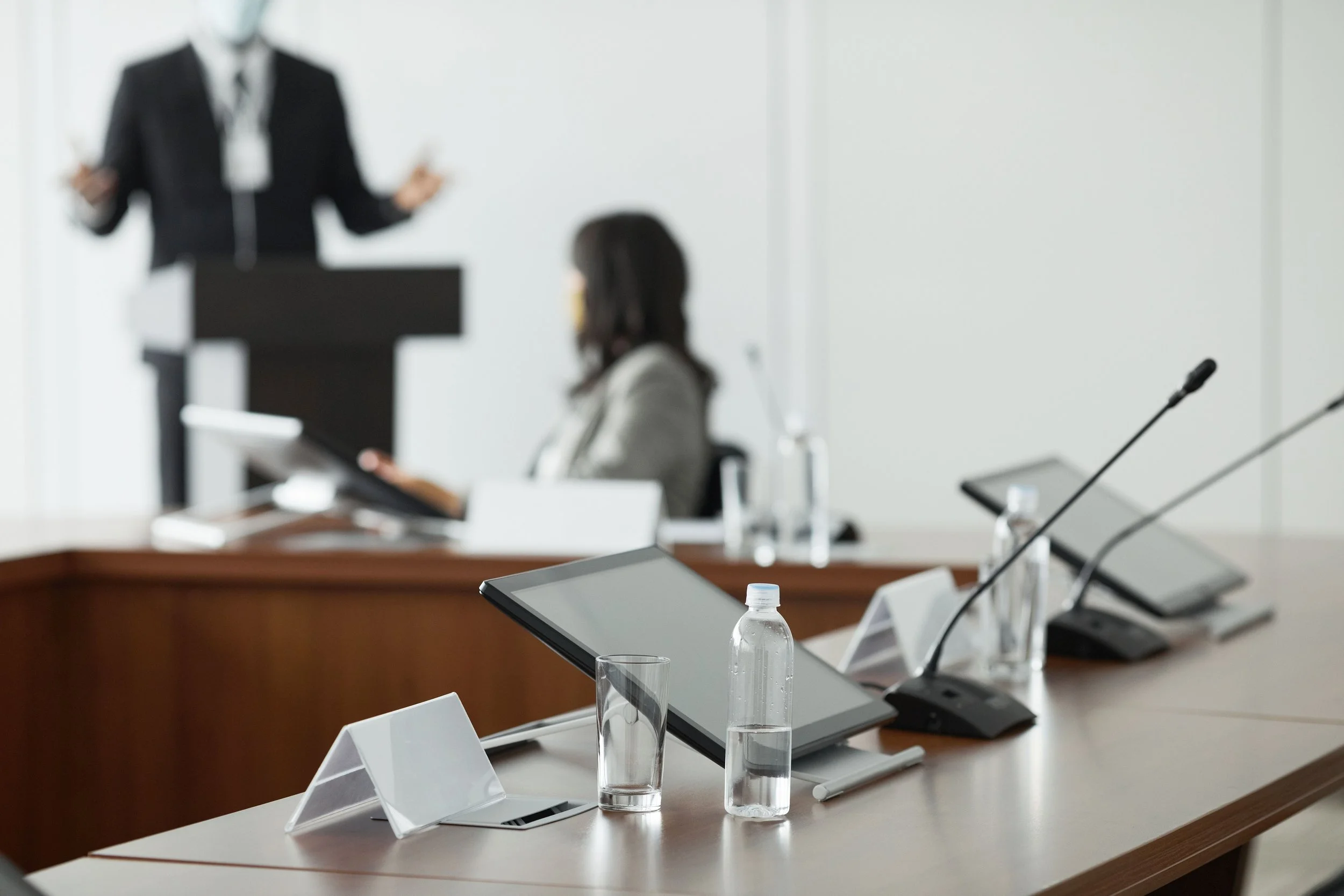 A blurred image of a conference room with a person giving a presentation at a podium in the background. The foreground shows a table with microphones, tablets, and water bottles.
