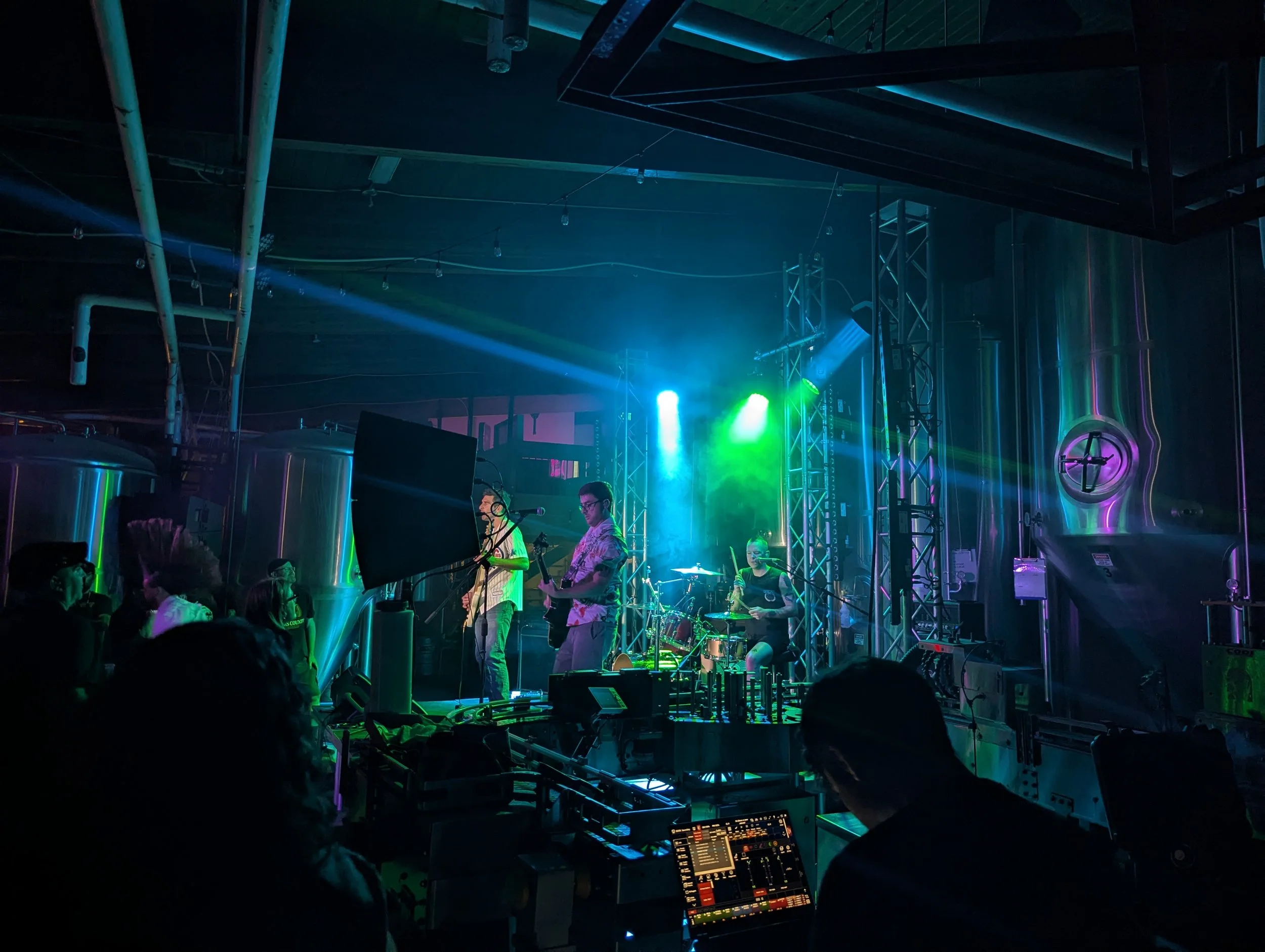 An indoor concert or nightclub scene with a band performing on stage, colorful stage lights, and an audience. Technical equipment and a DJ console are visible in the foreground.