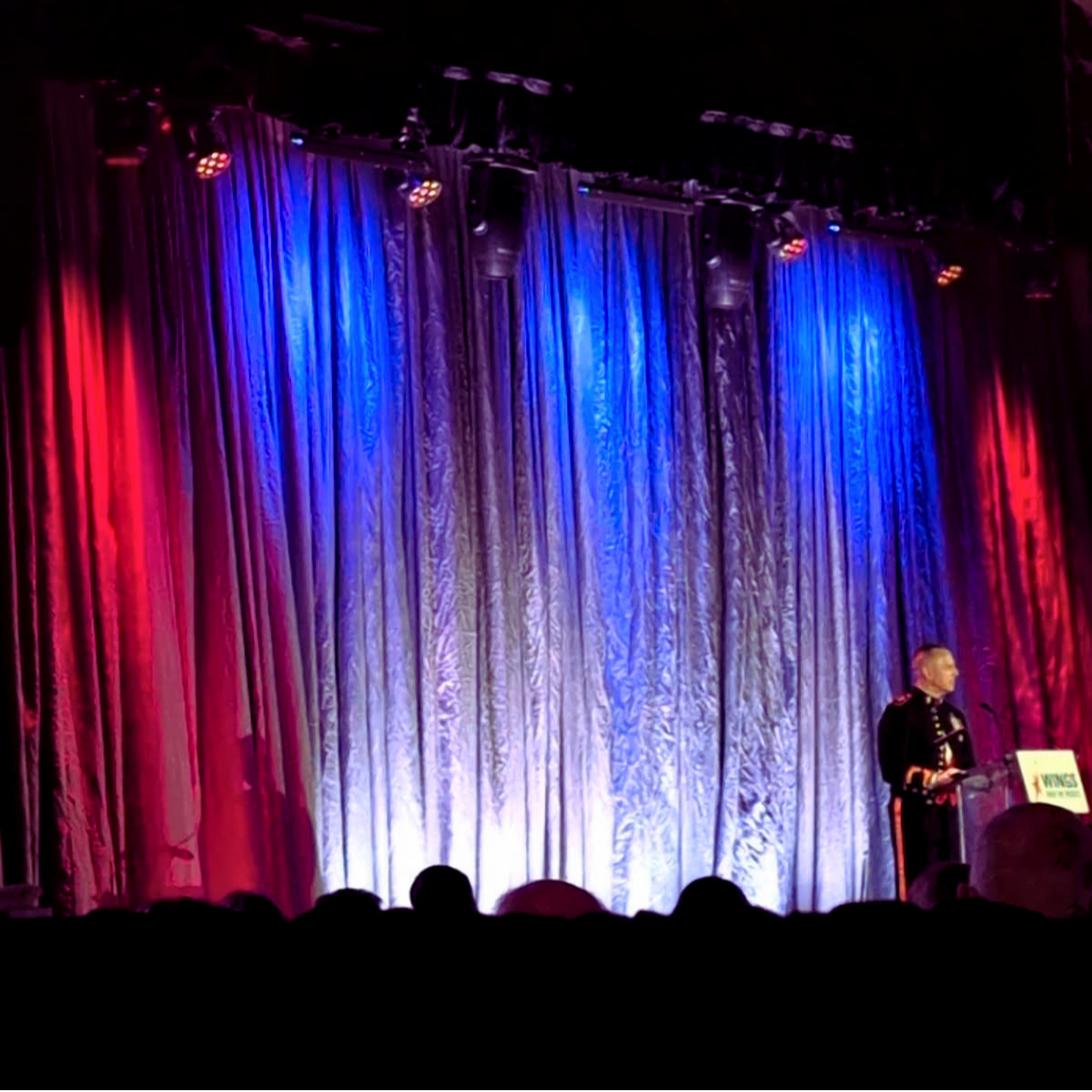 Stage with colorful curtains illuminated by red, blue, and purple lights. A person is standing at a podium on the right side.
