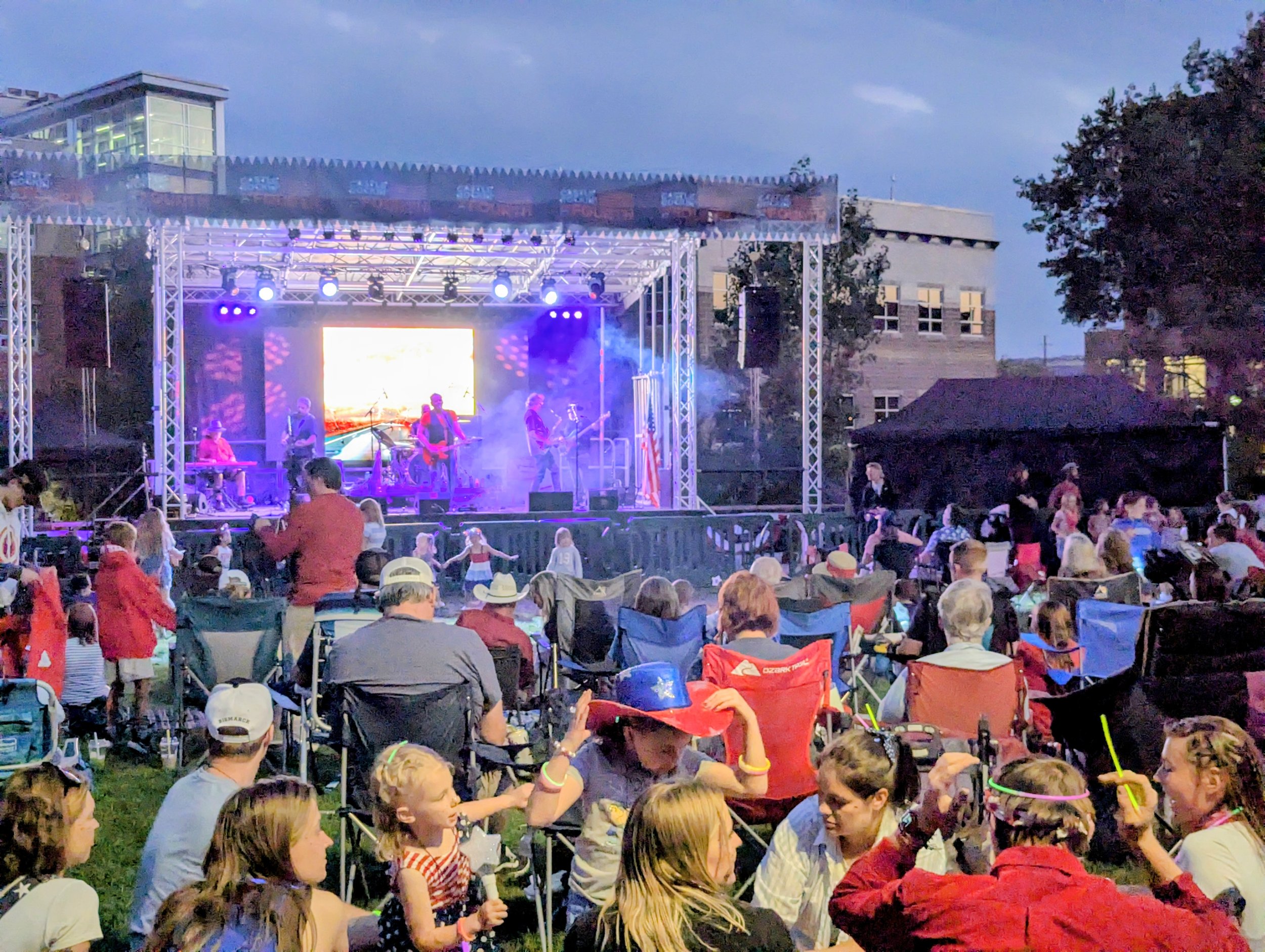 An outdoor concert at dusk with a stage and a band performing. Audience members, including children and adults, are seated in chairs and on the grass, some wearing glow accessories. The scene includes trees and buildings in the background.