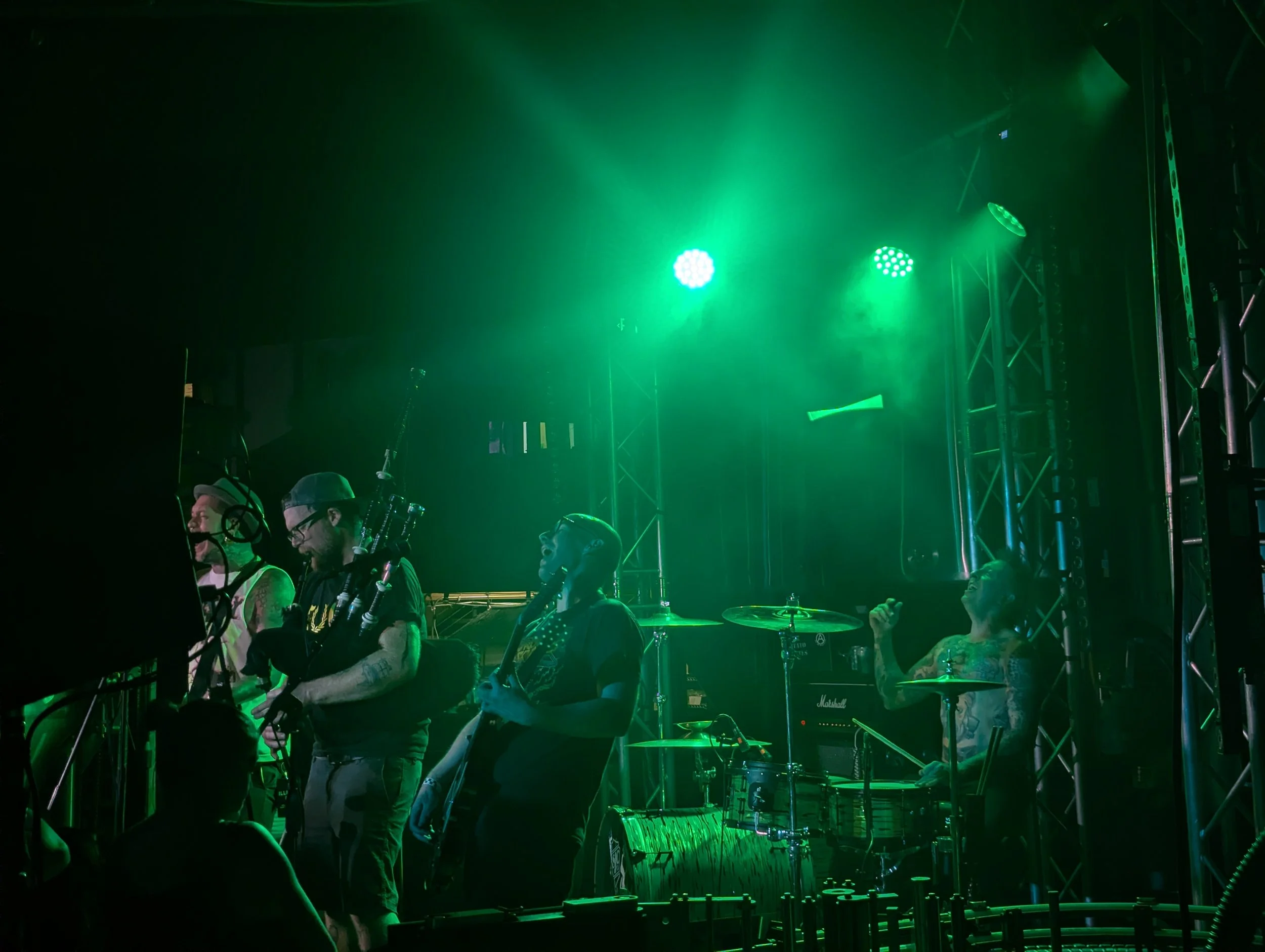 A group of musicians with percussion instruments on stage, illuminated by green stage lights, with equipment and structures visible in the background.