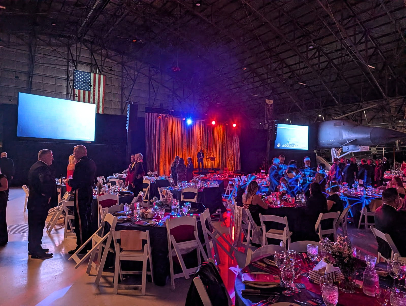 Indoor event space with tables set for a formal dinner, decorated with flowers and candles. A stage with a person speaking stands against a golden curtain, with colorful lights overhead. An American flag hangs on the wall, and there is a military jet