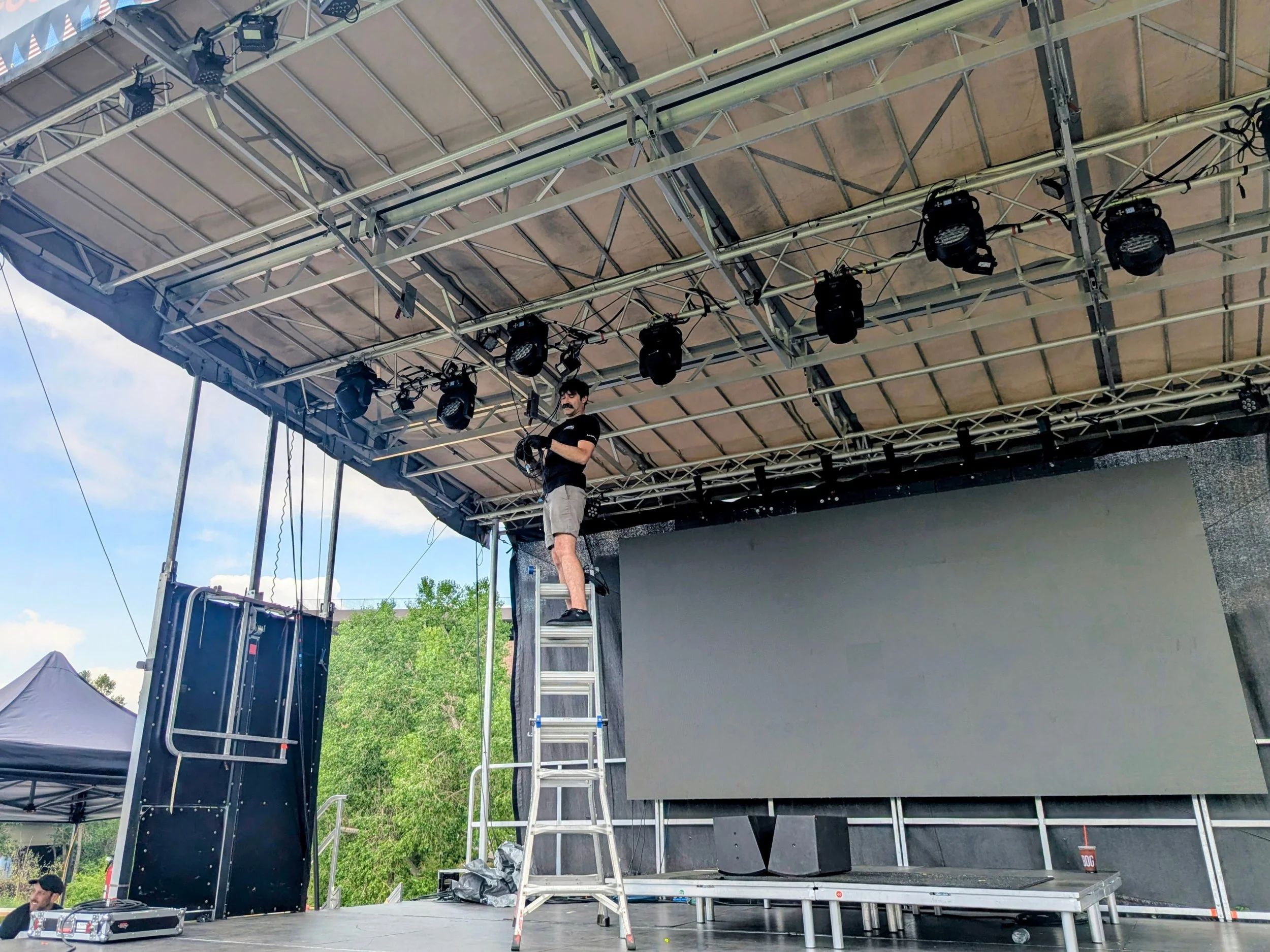 A person standing on a ladder working on a stage rigging system with spotlights and movers hung outdoors during daytime with trees and tents visible in the background.