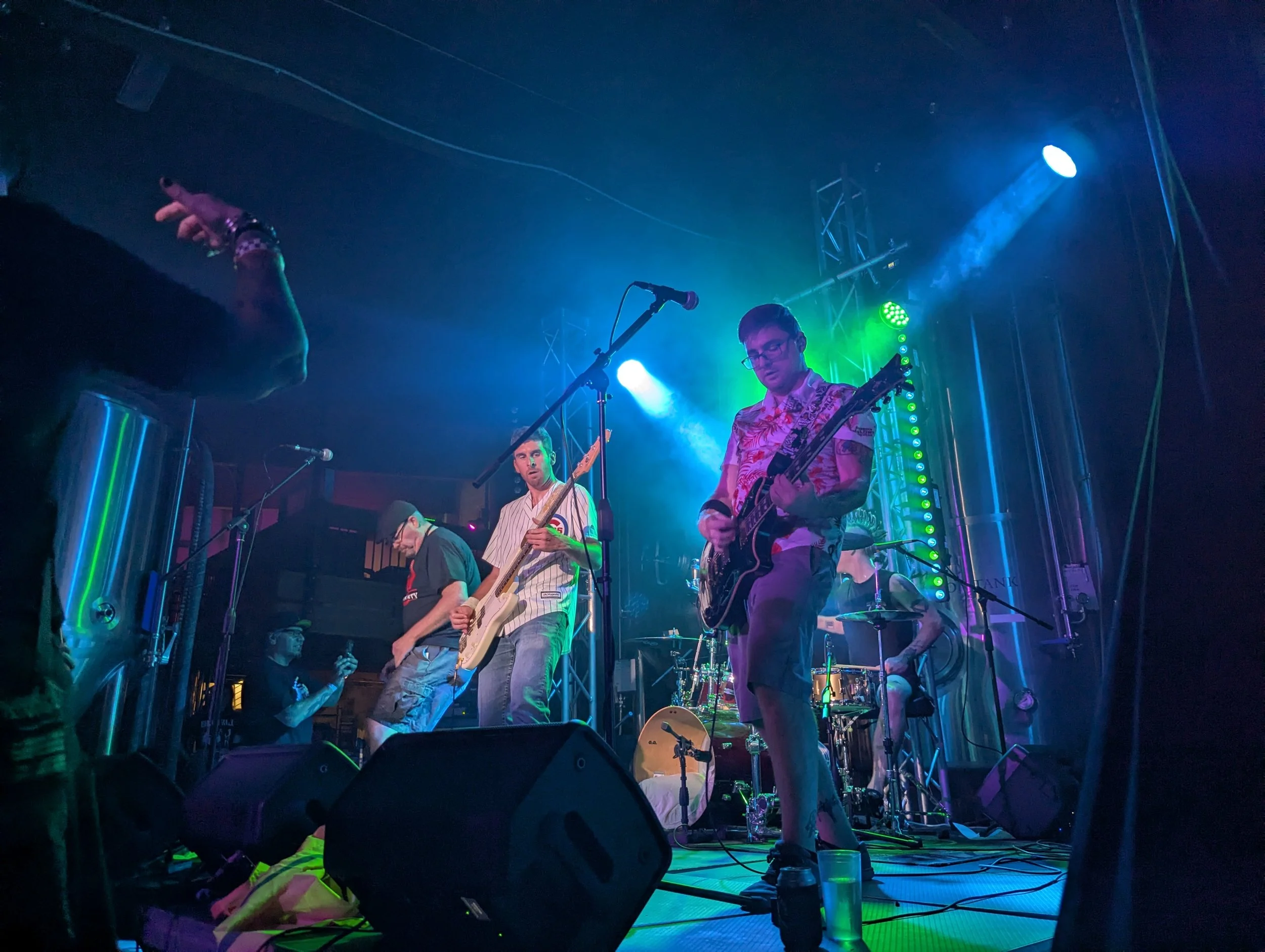A band performing live on stage with colorful lighting, including two guitar players, a bassist, and a keyboardist, with audience members visible in the foreground.