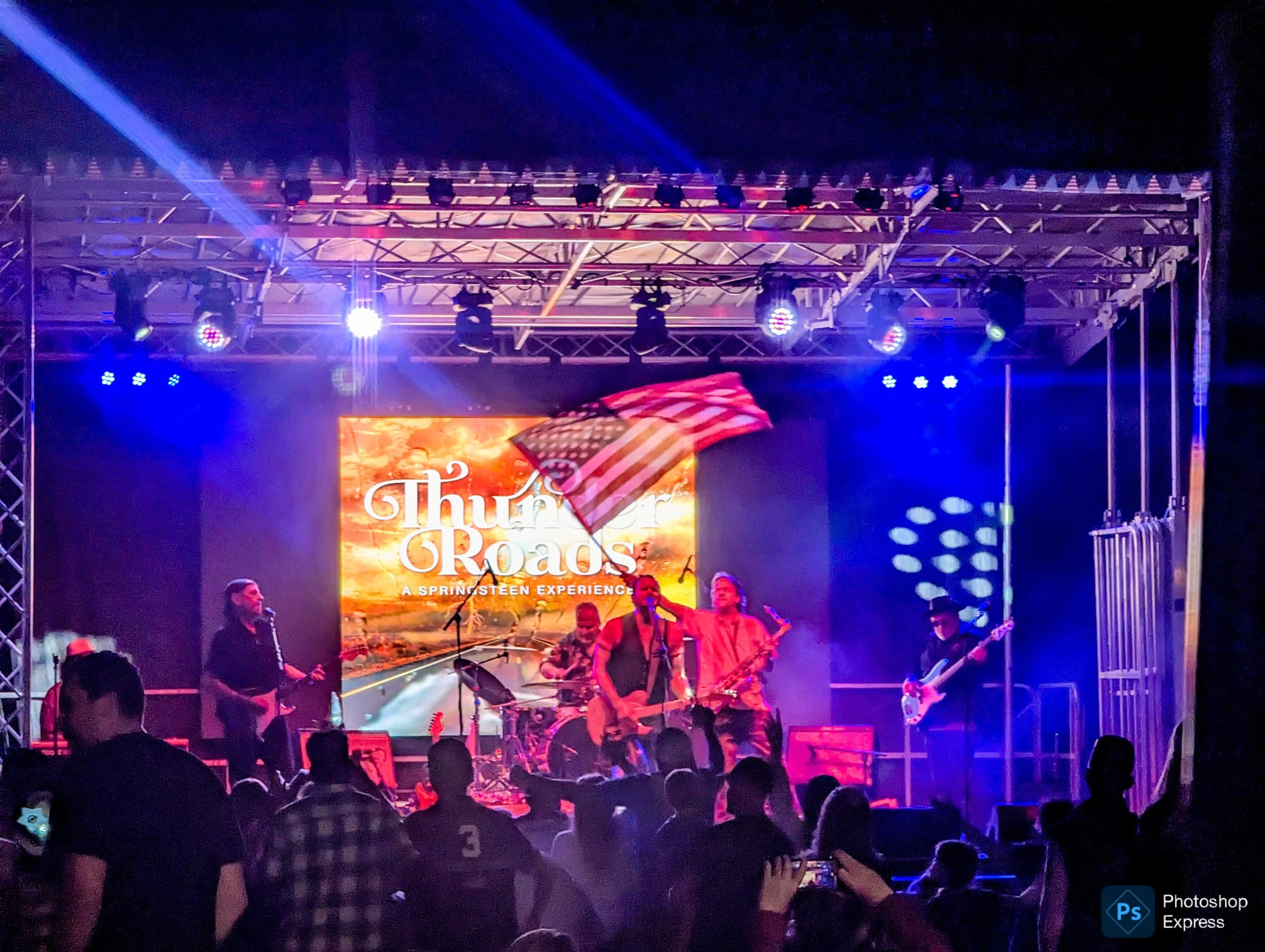 Stage with band performing under colorful lighting, large screen displaying the text "Thumper G" and a flag design, audience in the foreground.