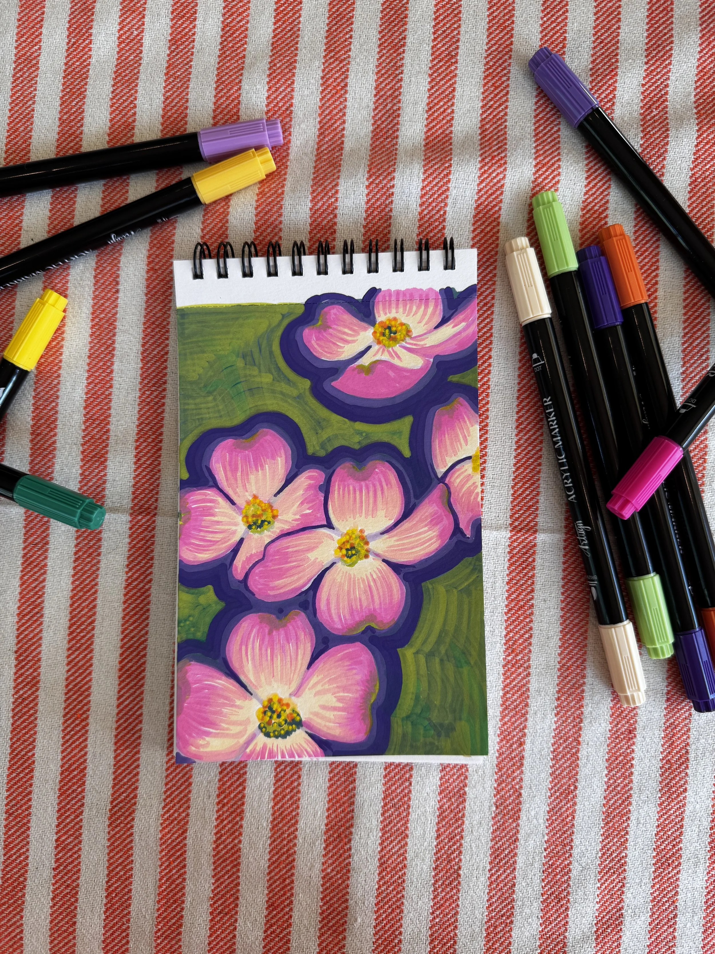 A small spiral-bound notepad with a colorful floral painting of pink flowers and green leaves is placed on a red and white striped fabric surface. Surrounding the notepad are several markers with colored caps, including purple, yellow, beige, orange, green, lavender, and pink.