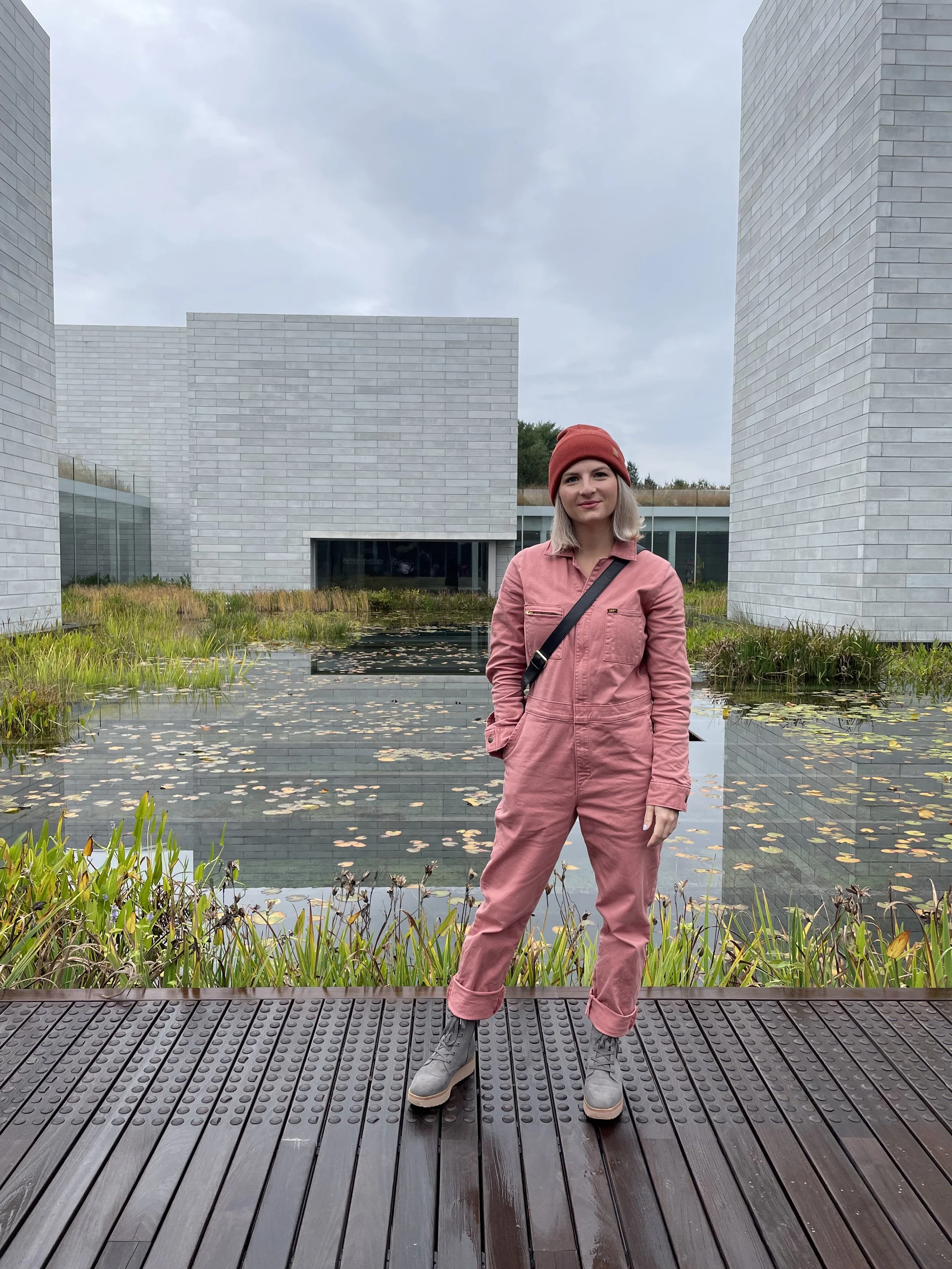 A woman standing on a wooden walkway by a pond with lily pads, in front of modern white brick buildings, wearing a pink jumpsuit, gray boots, and a red beanie.