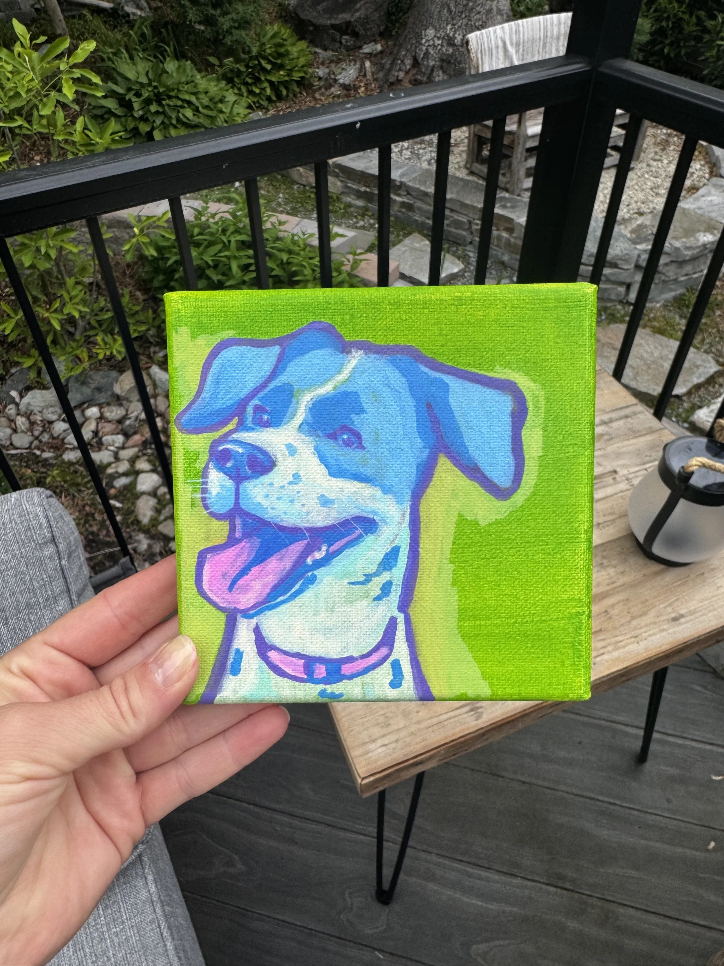 A hand holding a colorful painting of a dog on canvas with a bright green background, outdoors on a wooden deck with greenery and a stone step in the background.