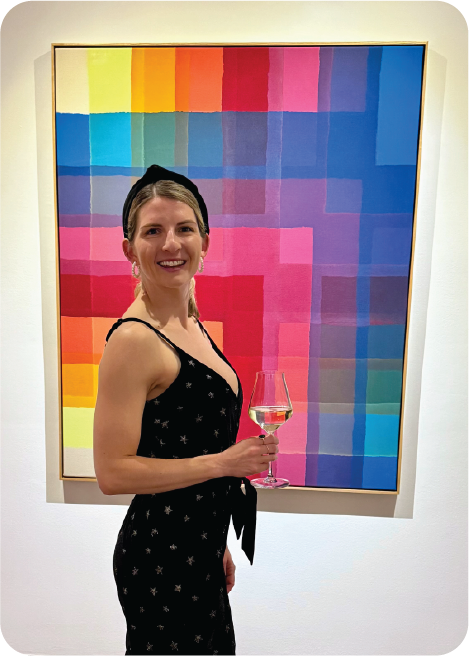 A woman in a black dress with a star pattern holding a glass of wine at an art gallery with a colorful, geometric abstract painting in the background.