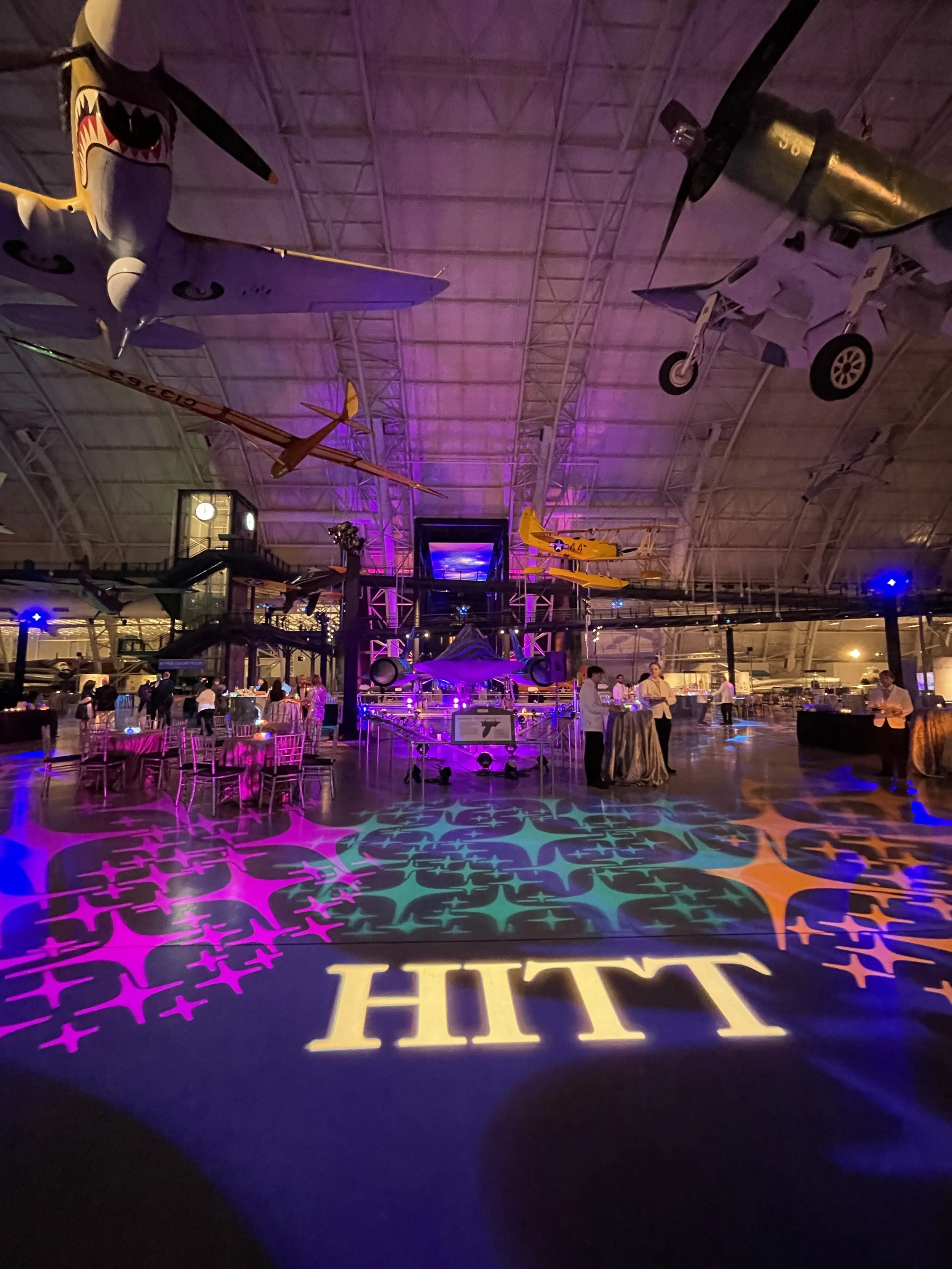 An indoor exhibit with hanging model airplanes and a stage area illuminated by purple and pink lighting, with a large sign on the floor reading 'THU' and reflective patterns projected onto the floor.