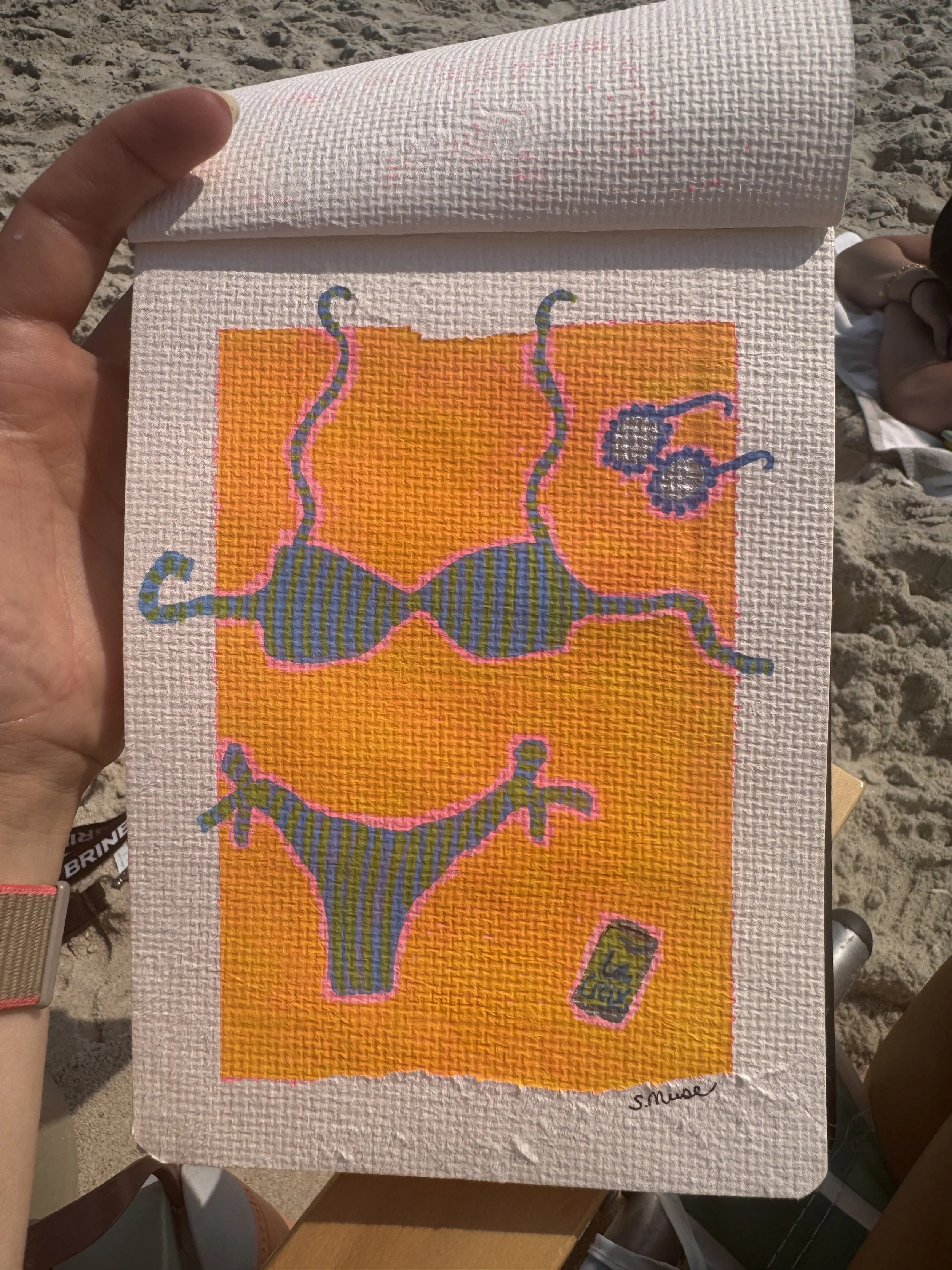 Cross-stitch artwork of a stylized blue bee with yellow and orange details, set against an orange background. The artwork is signed 'S. Muse' in the bottom right corner and appears to be held outdoors on a sandy surface.