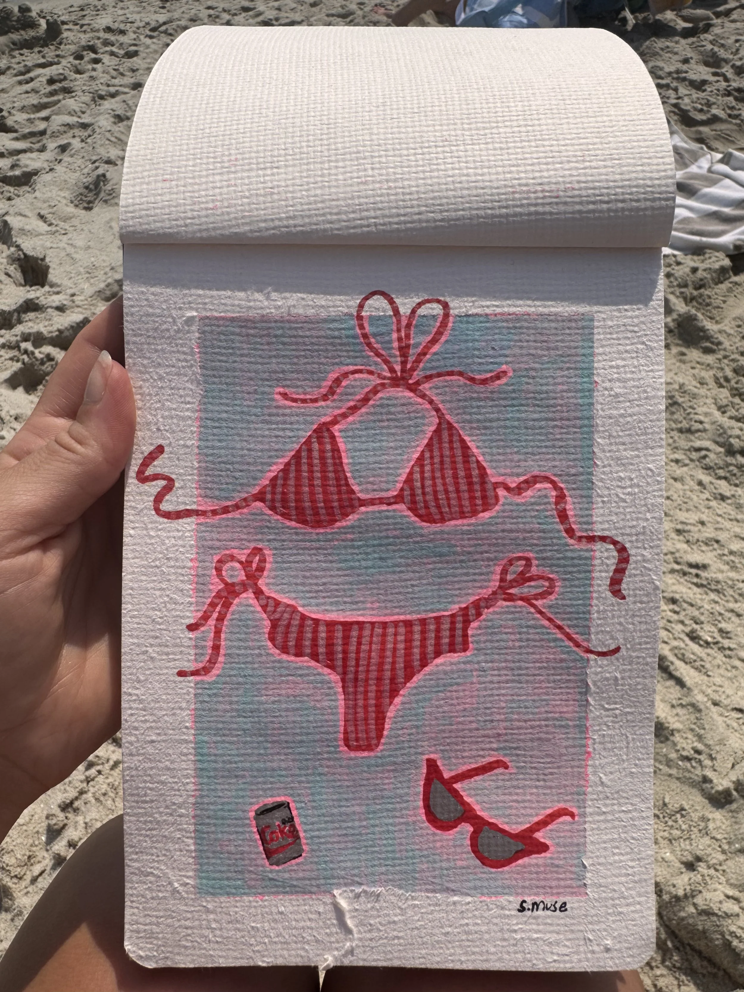 Drawing of a bikini with cherries, a soda can, and heart shapes, signed by S. Muse, held against a sandy beach background.