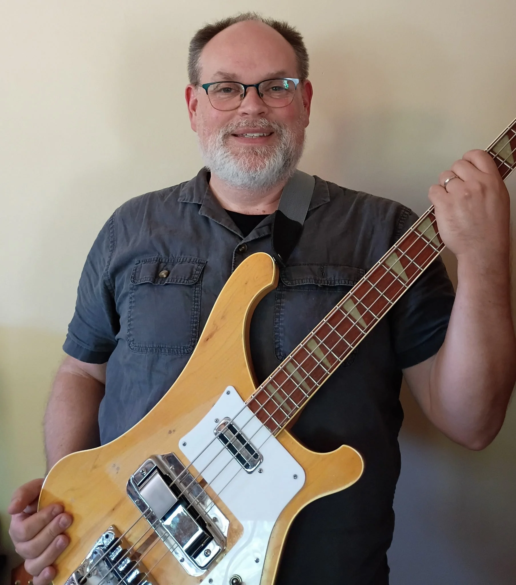 A man with glasses and a beard holding an electric guitar, smiling, against a plain wall background.