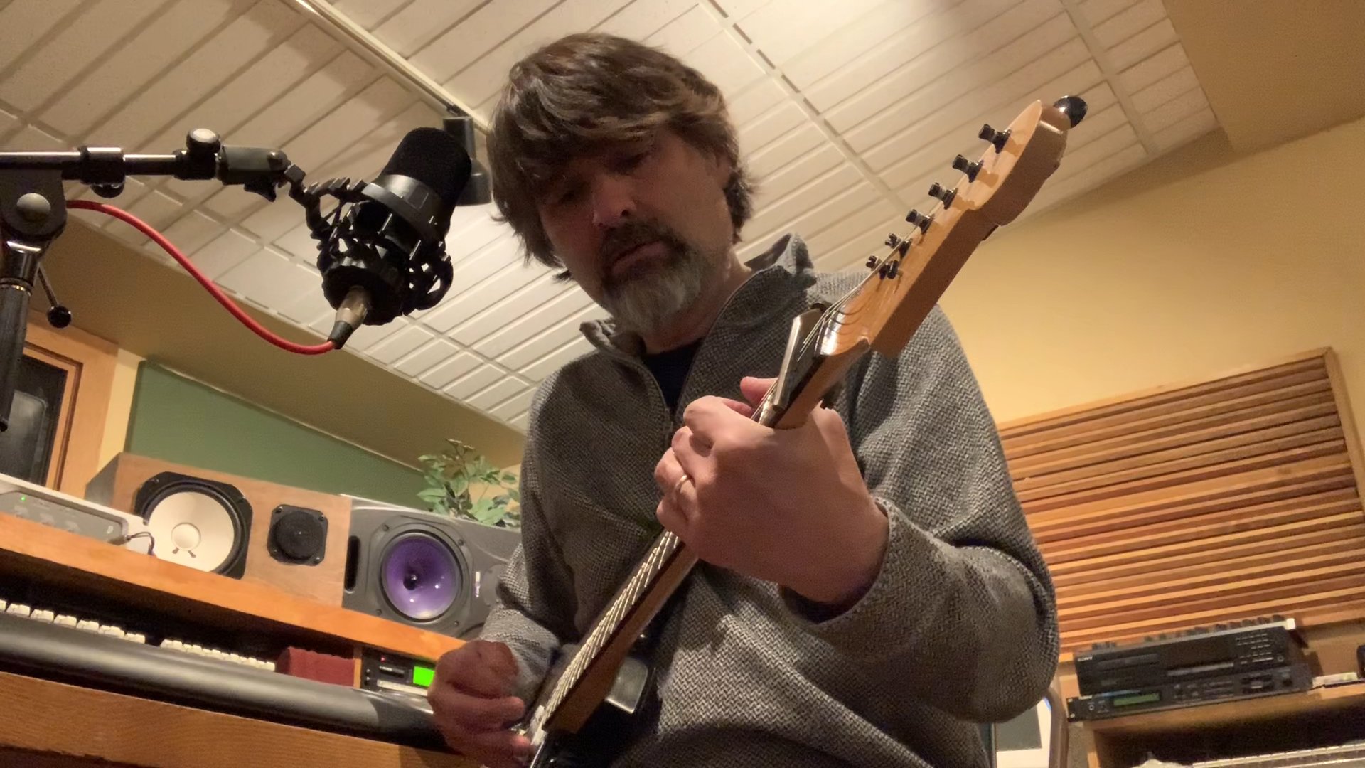 A man with brown hair and a beard playing an electric guitar in a recording studio. There is professional audio equipment, a microphone, and wood paneling in the background.