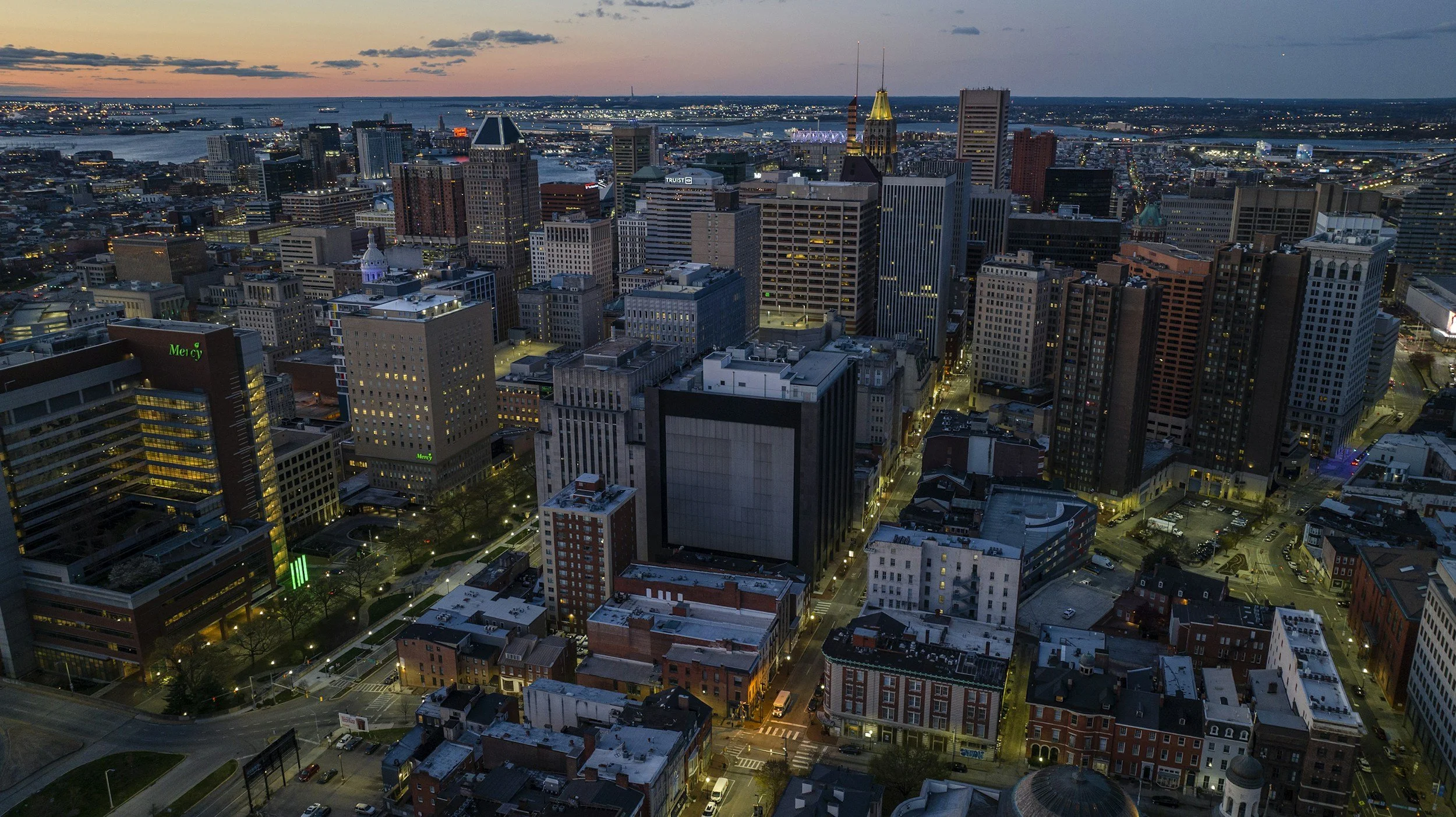 Aerial view of a city skyline at dusk with tall skyscrapers, a river in the background, and city lights illuminating the streets below.