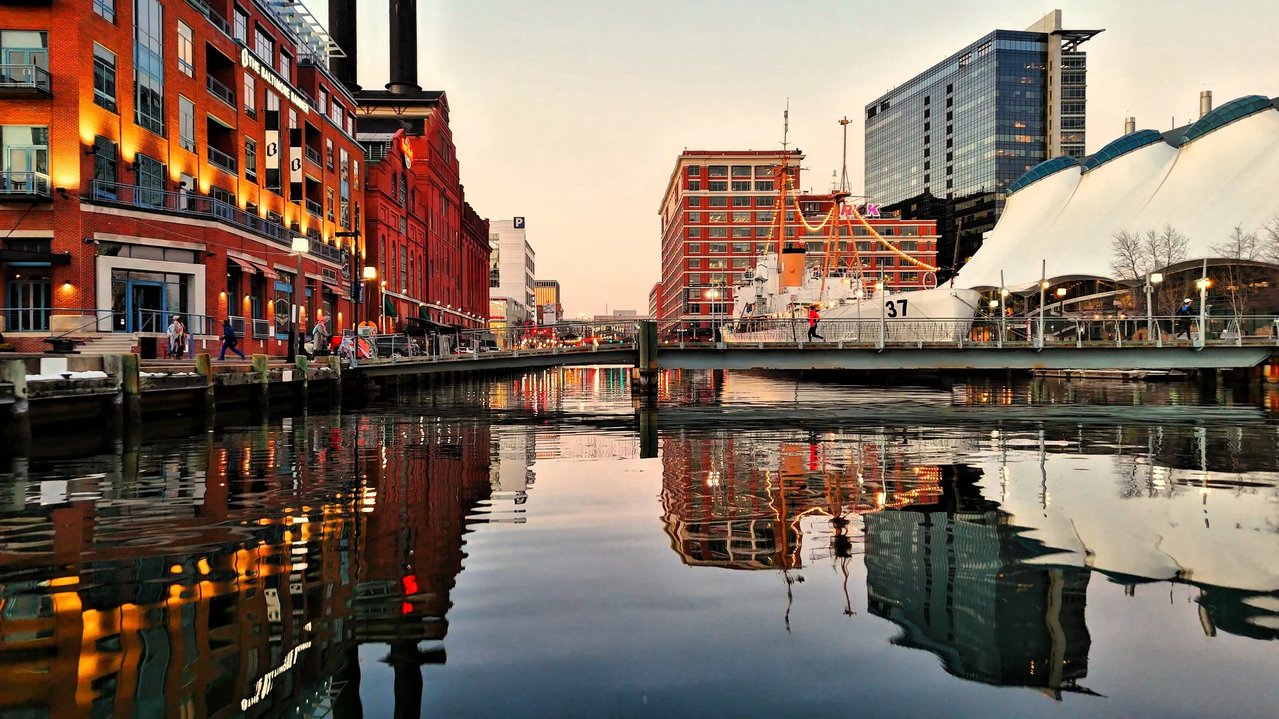 Cityscape with buildings, a bridge over water, and a boat with string lights, reflecting in the water during sunset.