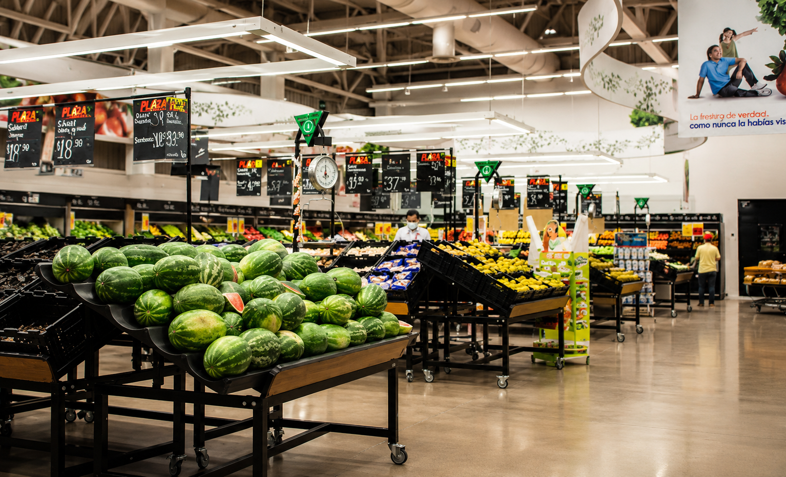 Carpa de supermercado con stand de sandías y otras frutas. Personas comprando, escaleras con productos, carteles con precios, decoraciones verdes en el techo.