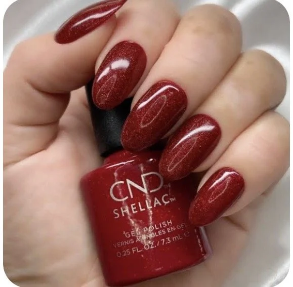 Close-up of a hand with long, almond-shaped nails painted in a glittery red nail polish, holding a small bottle of CND Shellac gel polish in matching red color.