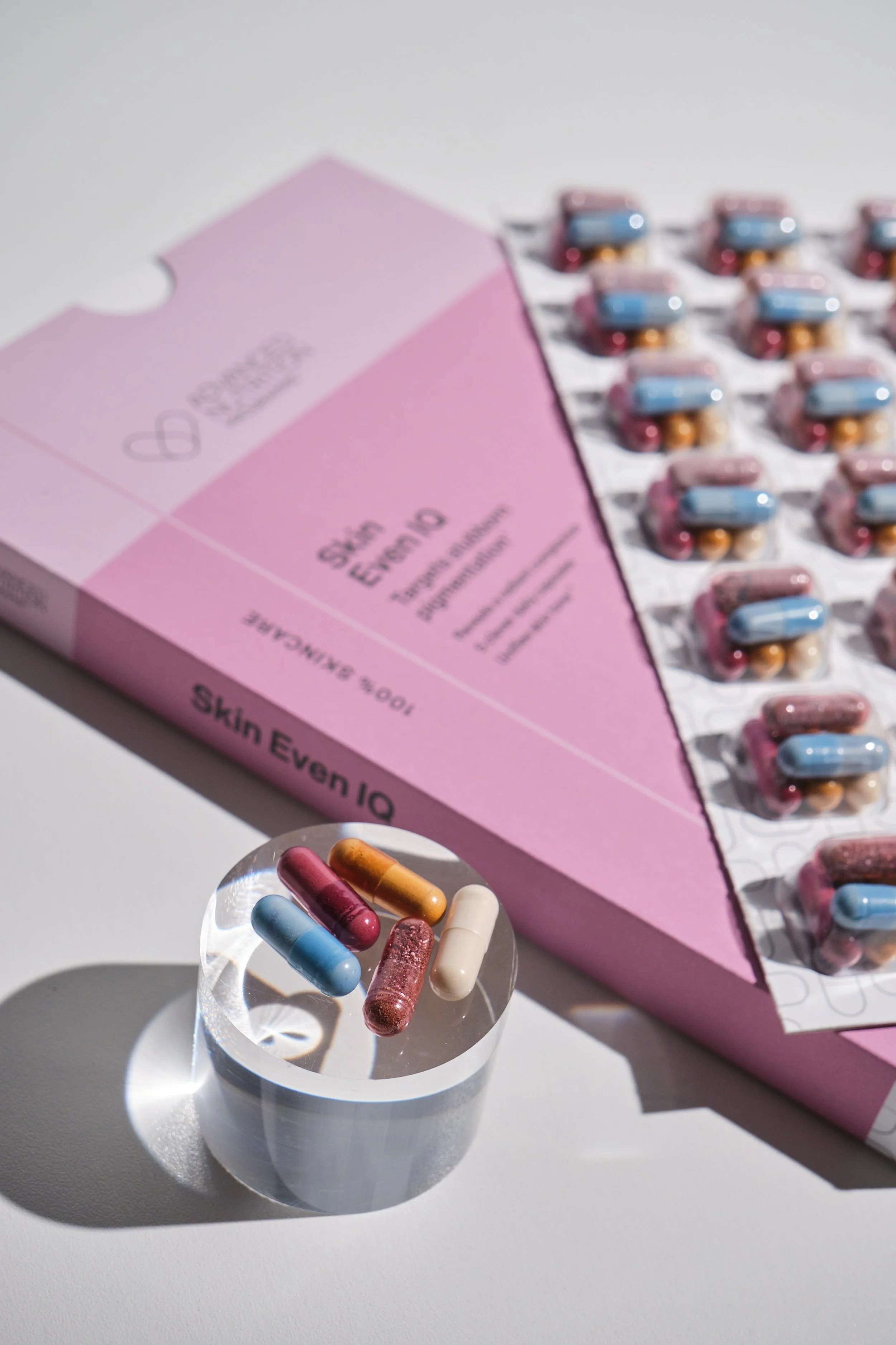 Close-up of capsules in a small glass container, with a pink box labeled 'Skin Event IQ' containing blister packs of capsules in the background.