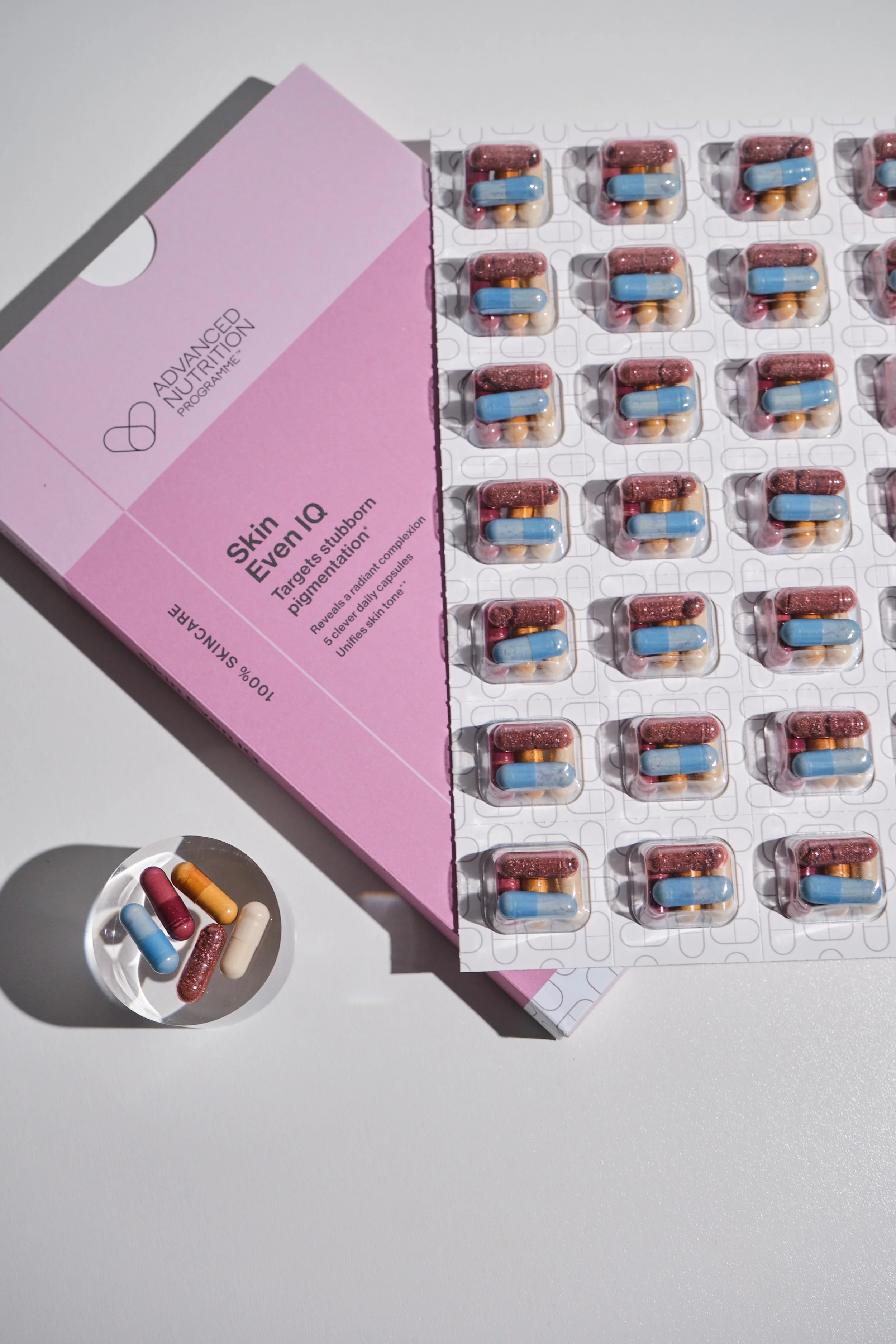 A pink box labeled 'Advanced Nutrition Program' with the name 'Skin Even IQ' and description 'Targets stubborn pigmentation'. Next to it are blister packs of capsules with blue, pink, white, and glittery red colors, along with a small dish holding some capsules.