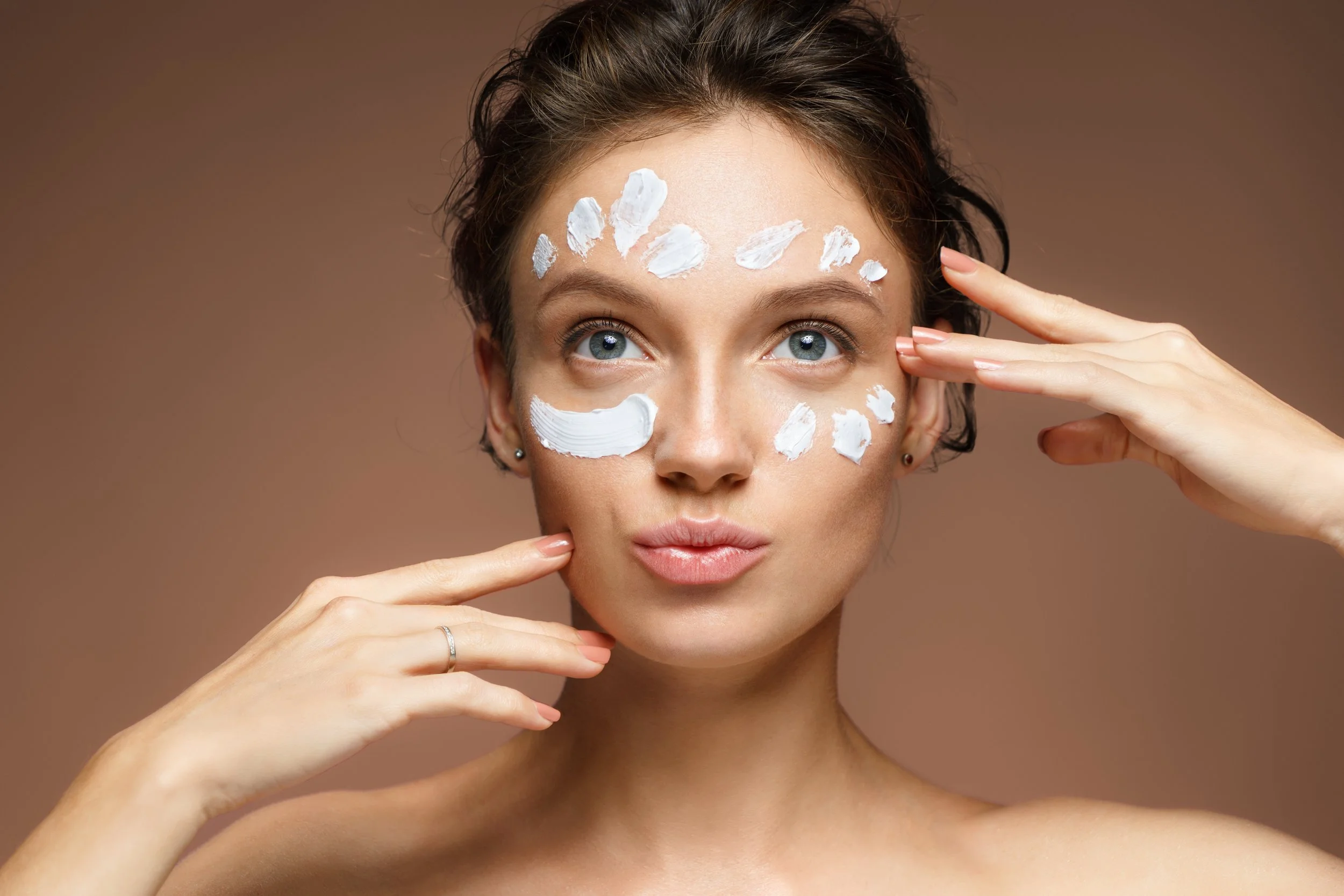 A woman applying white cream to her face, with some on her forehead, cheeks, and chin, while looking directly ahead.