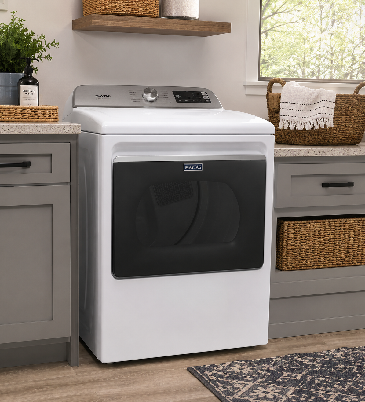 A Maytag front-loading washing machine in a laundry room with gray cabinets, a woven basket, a potted plant, and a window with a view of trees outside.